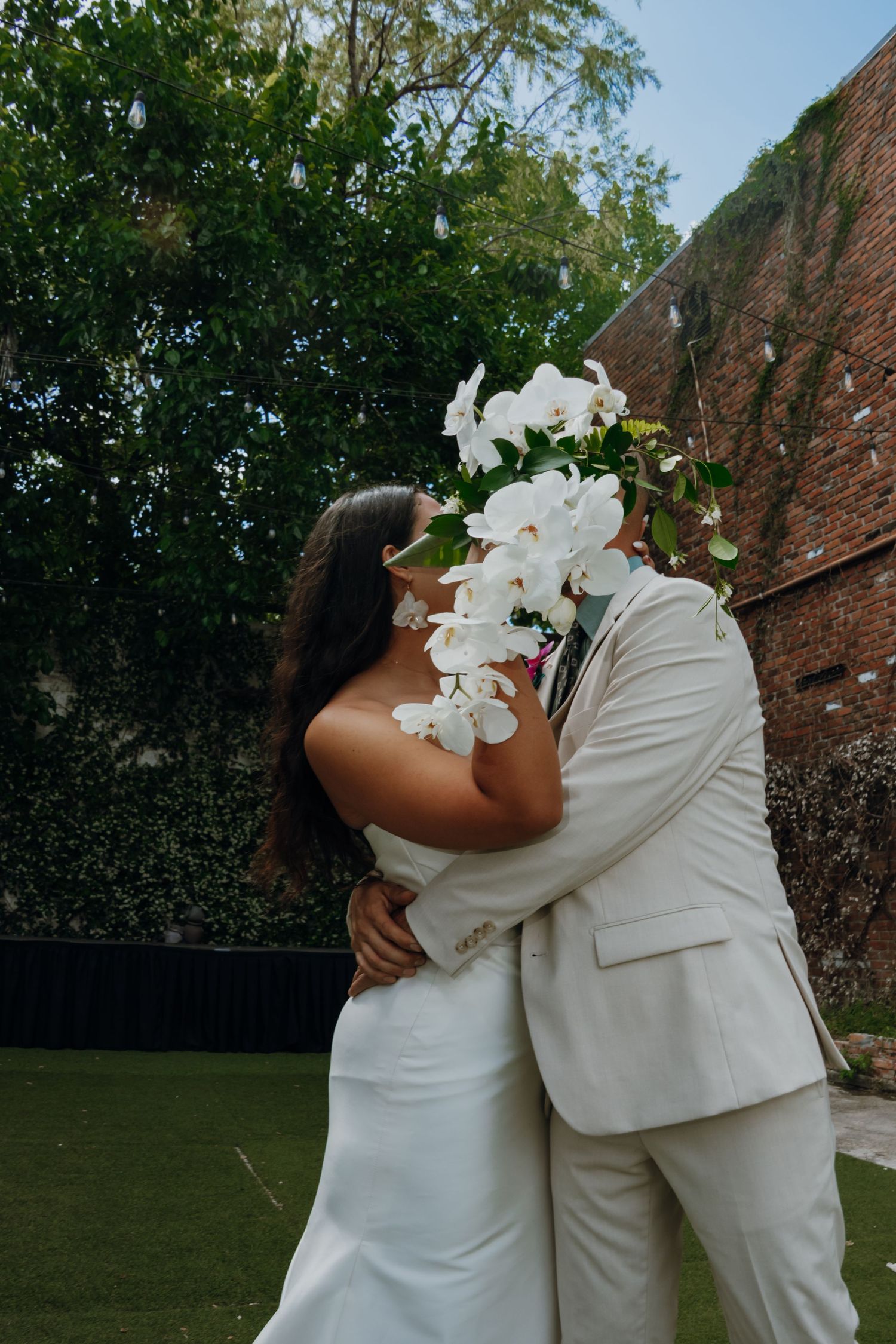 Bride and groom share a kiss hidden behind large white orchid bouquet in garden setting.