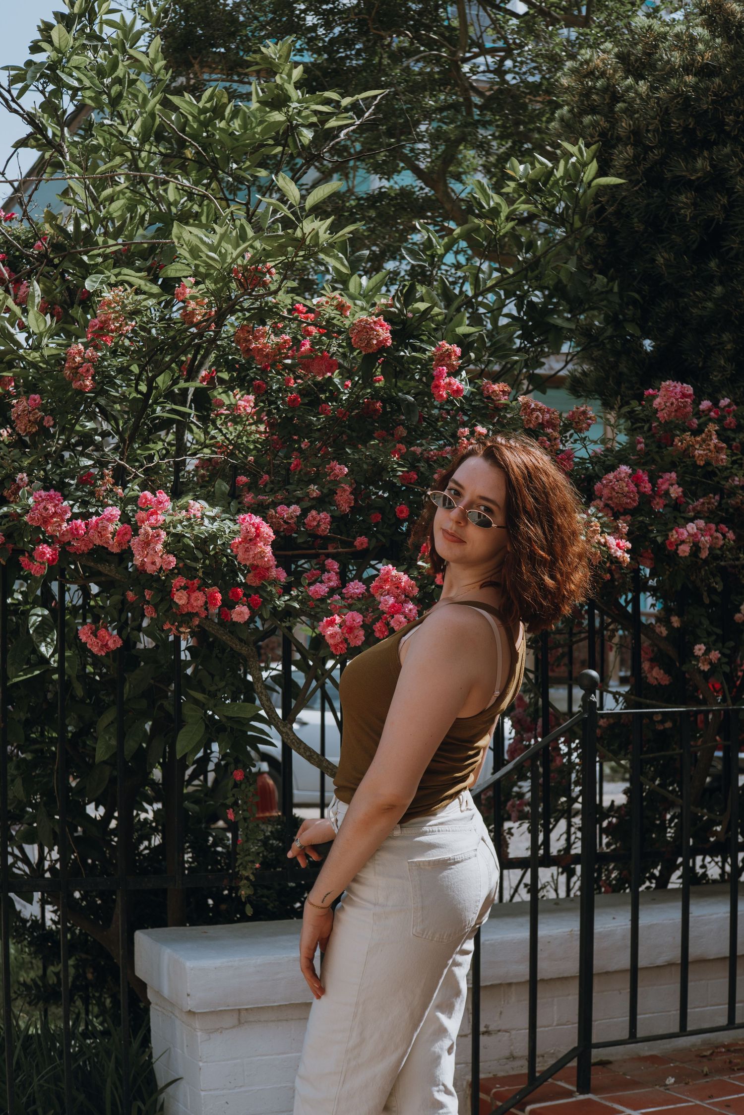 Young woman in olive tank top and white pants posing beside blooming pink rose bushes behind iron fence.