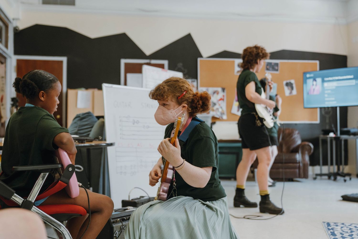 Redhead student plays ukulele seated in music classroom while classmate watches and teacher plays guitar. New Orleans, Louisiana, Mississipp