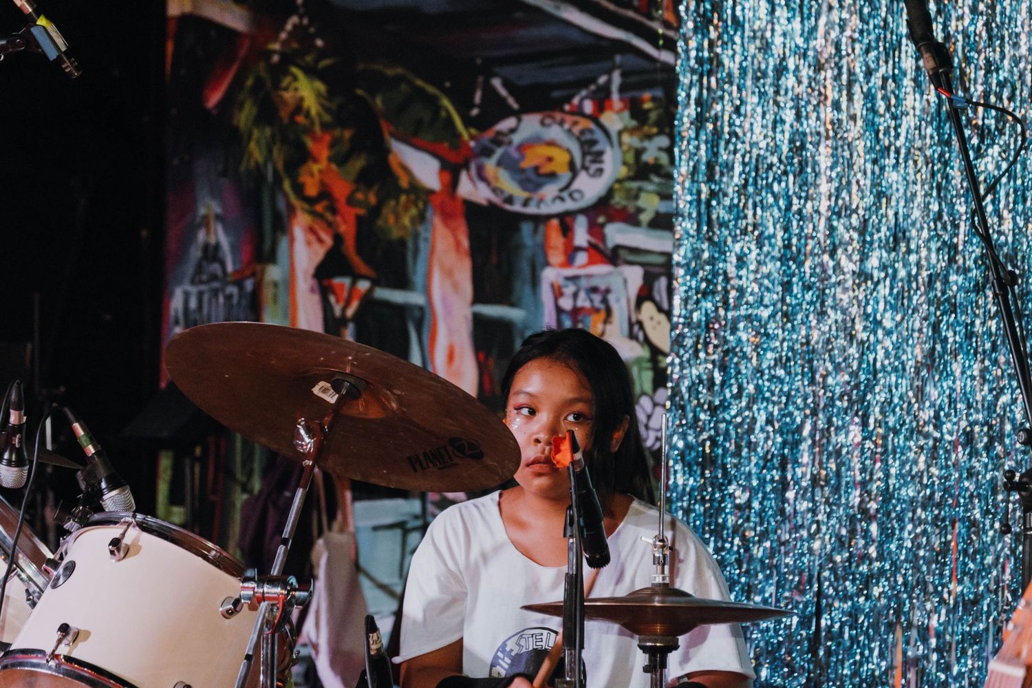 Young girl playing drums on stage with a sparkling blue curtain backdrop.