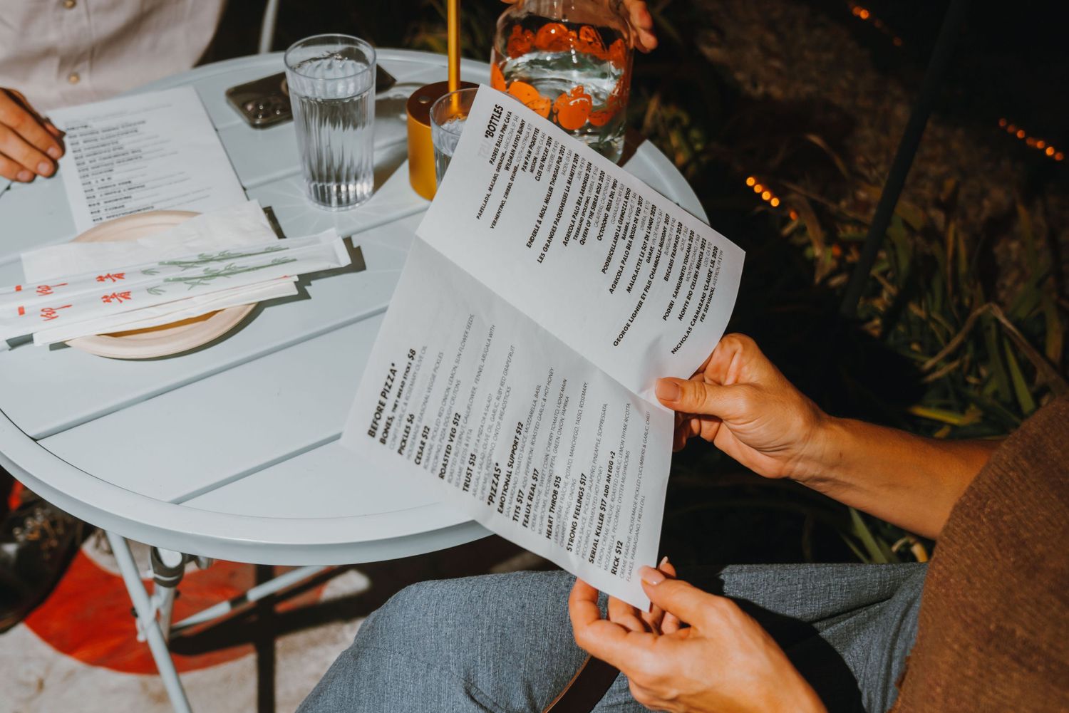 Two people reading restaurant menus at an outdoor white bistro table with drinks at night.