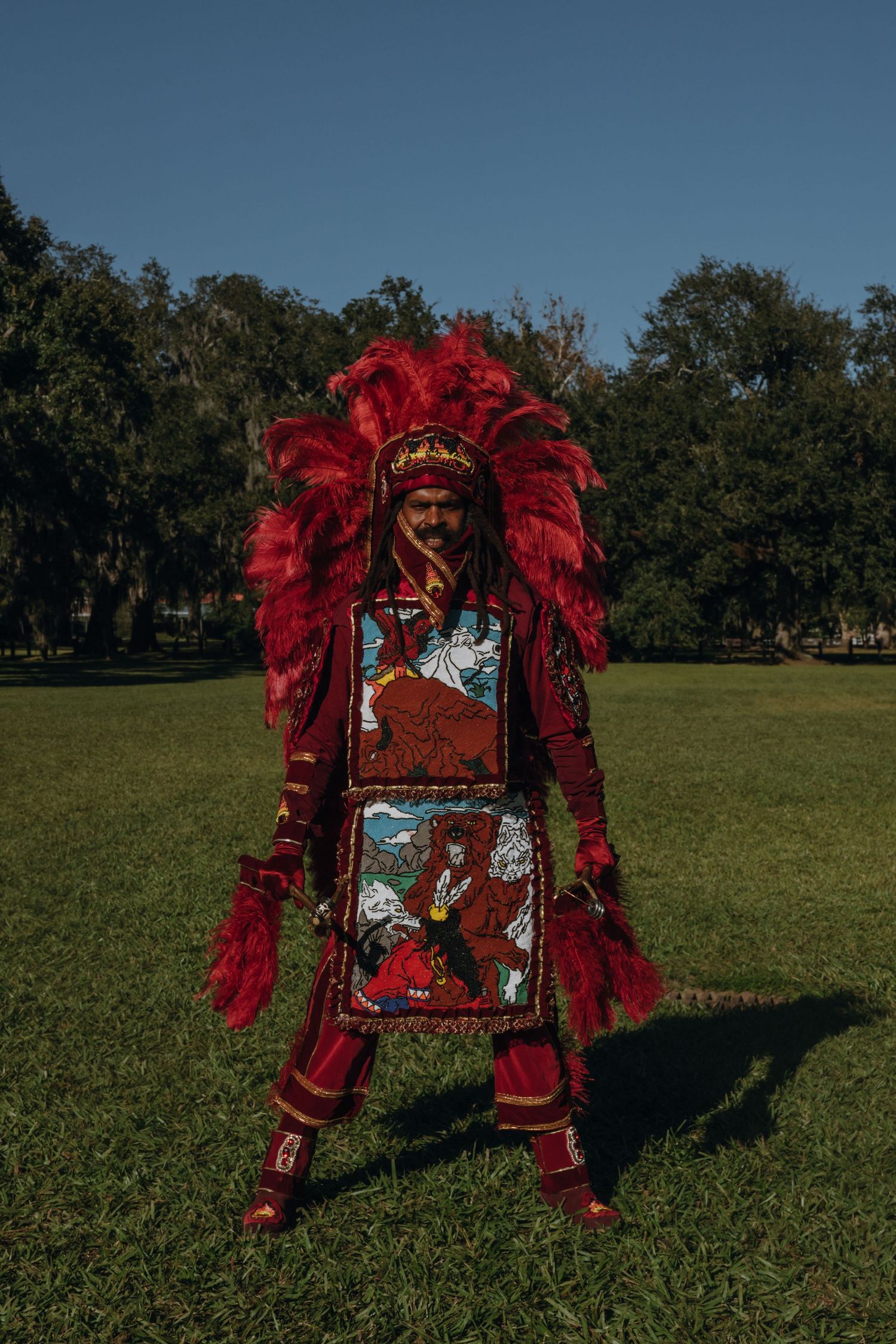 Mardi Gras Indian in red feathered crown and intricately beaded apron panels poses on green grass.