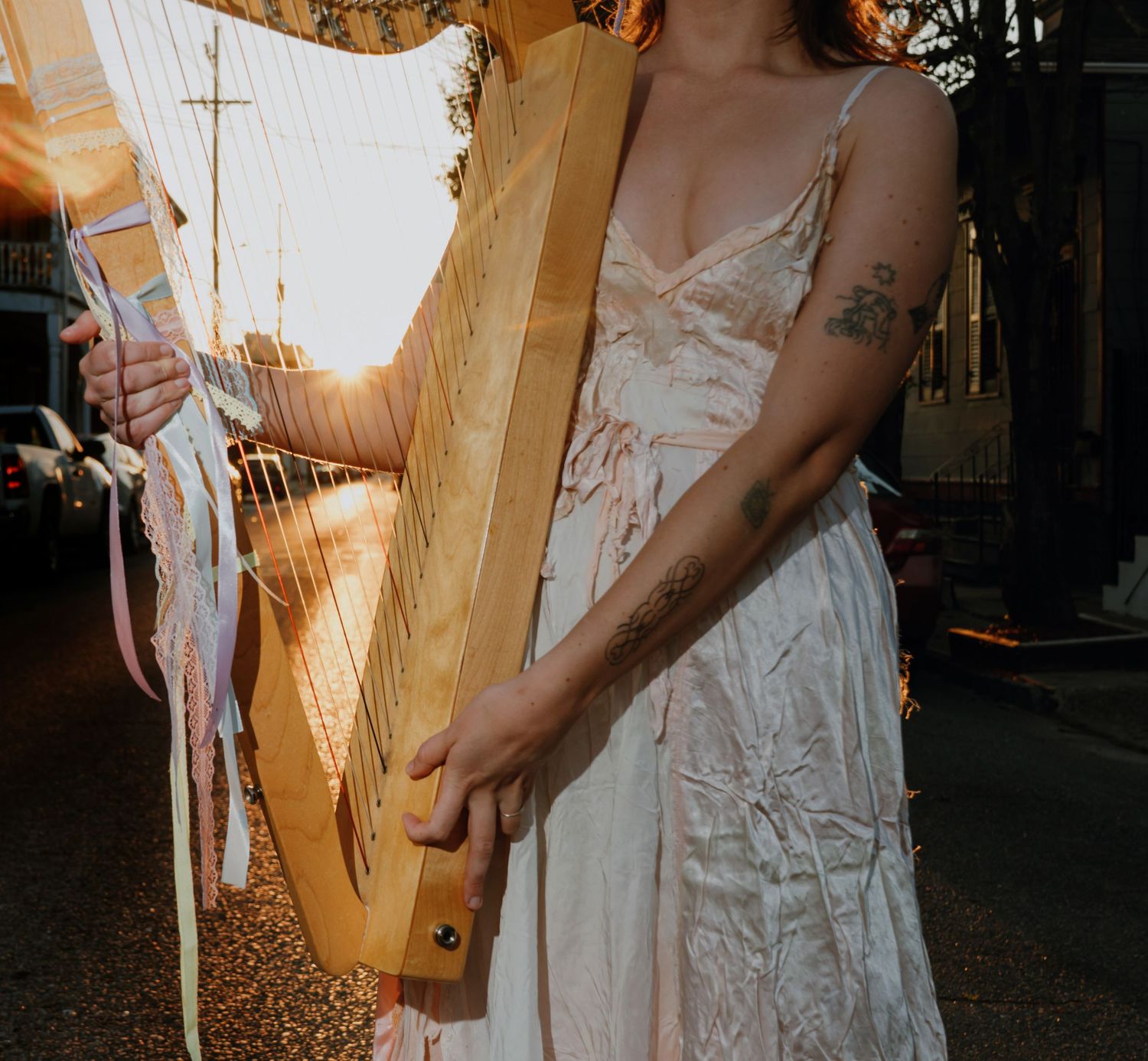 Woman in white lace dress playing wooden harp outdoors with warm sunset light glowing through strings.