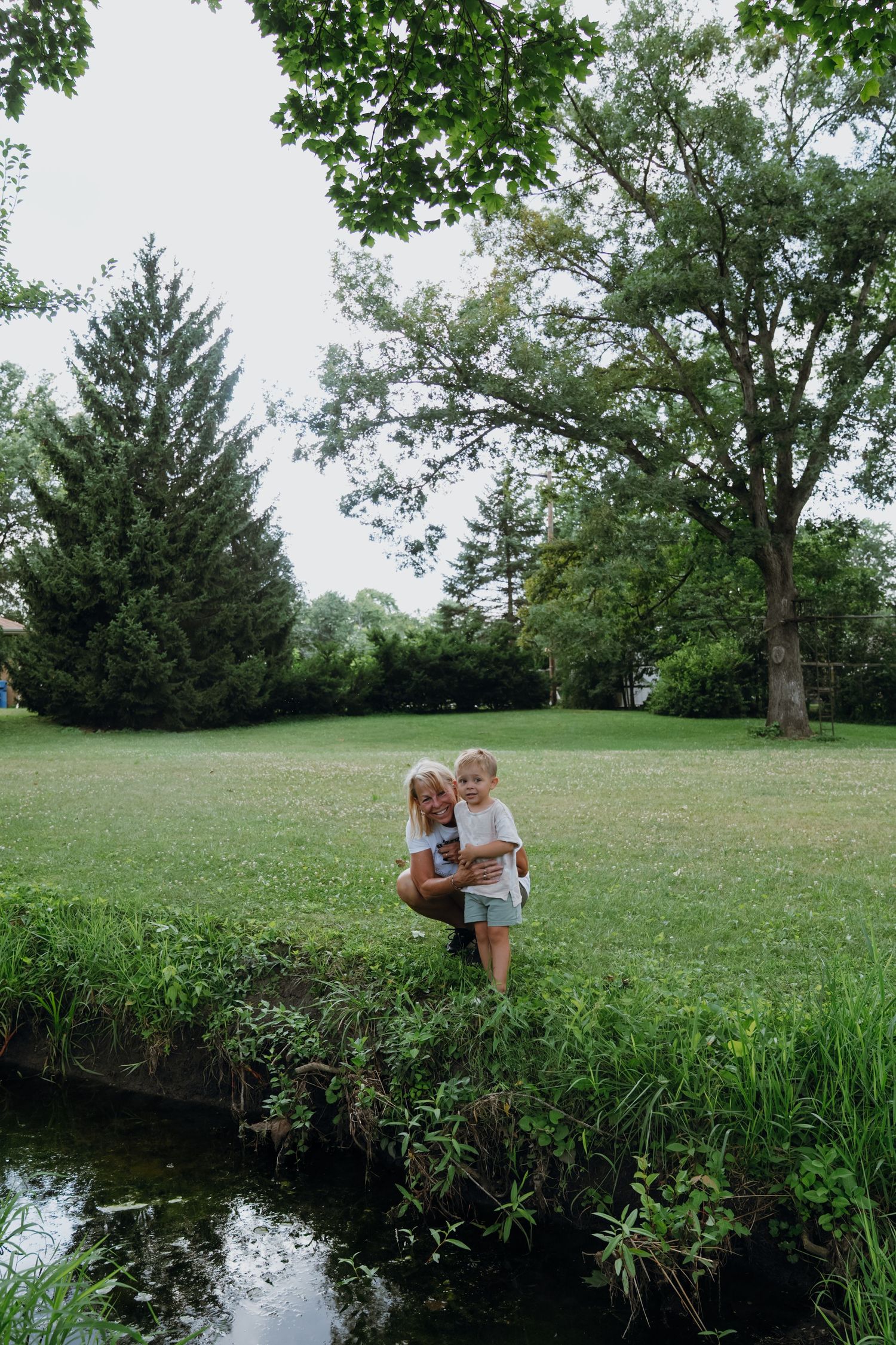 Mother crouching with toddler by a small stream in a lush green park setting.