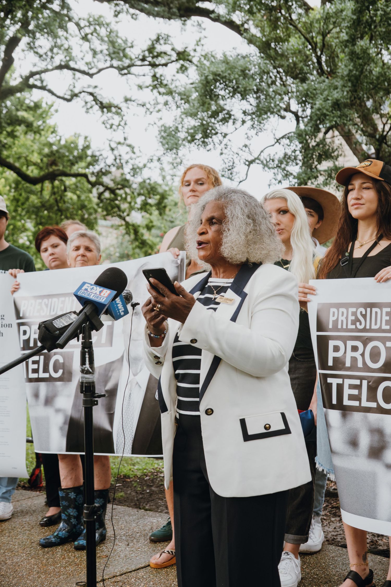 Woman speaks at outdoor press conference with microphones and protesters holding signs.