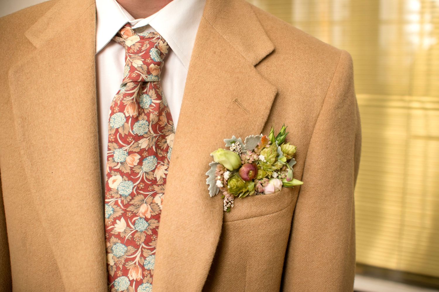 A man in a tan suit wearing a floral tie and decorative boutonniere with berries and greenery.
