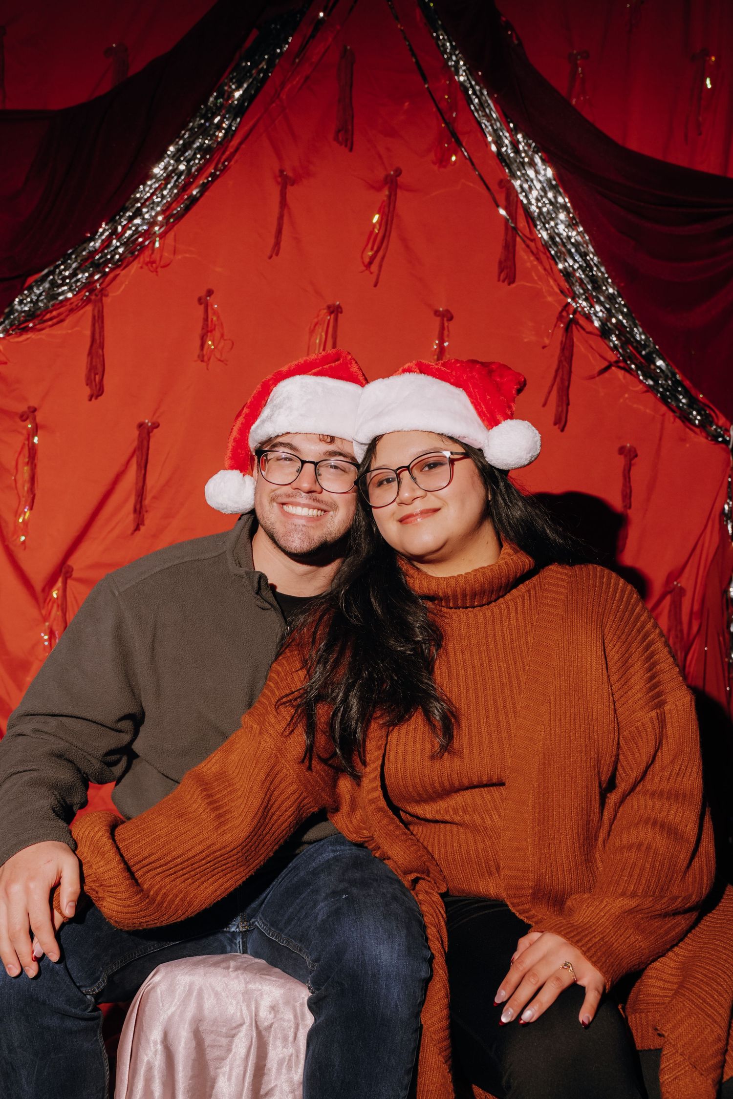 A smiling couple wearing Santa hats and glasses pose together in front of a festive red backdrop.