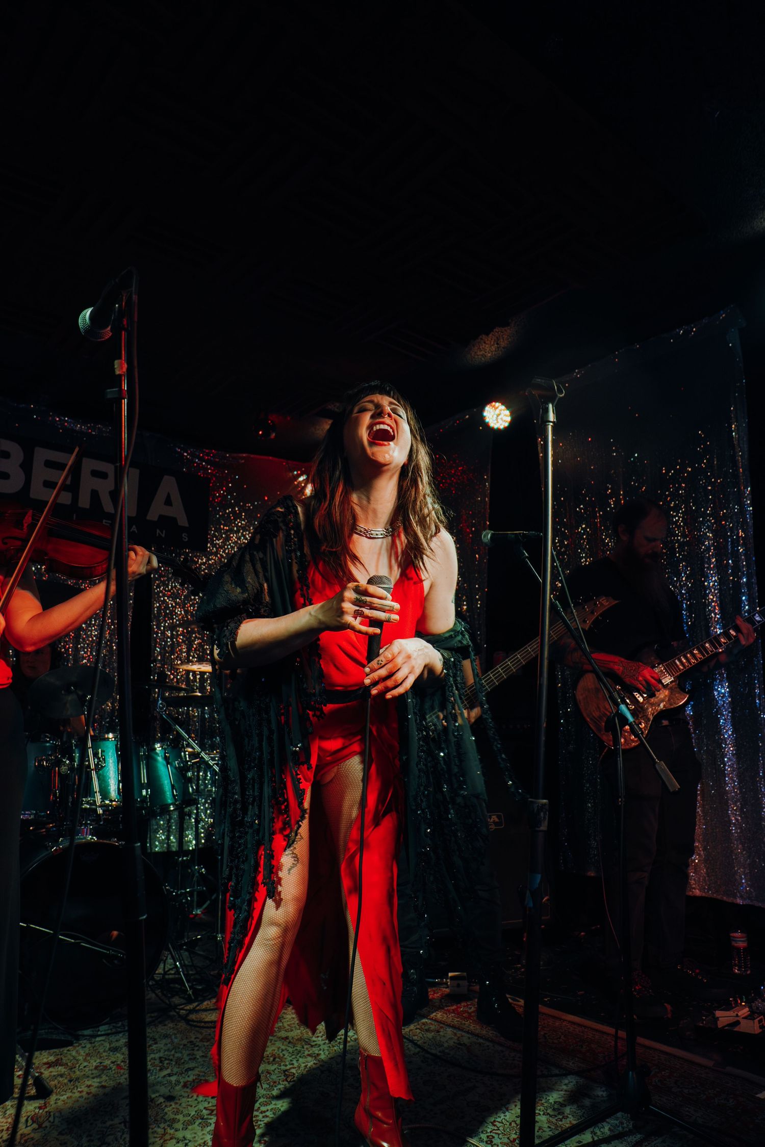 Female singer in red dress performing passionately on stage with band under colorful stage lights.