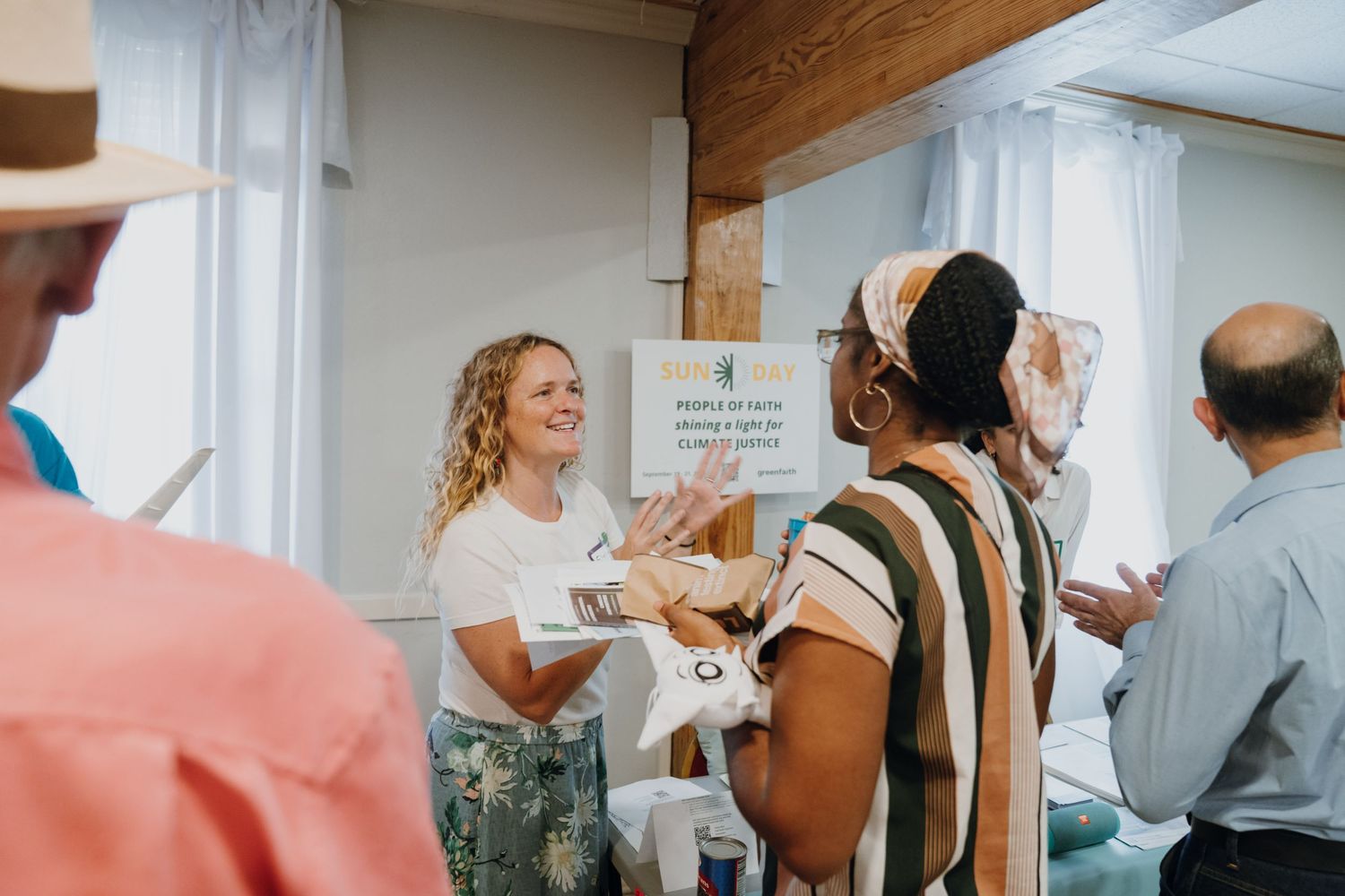 Woman in white shirt smiling and greeting attendees at an indoor community event.