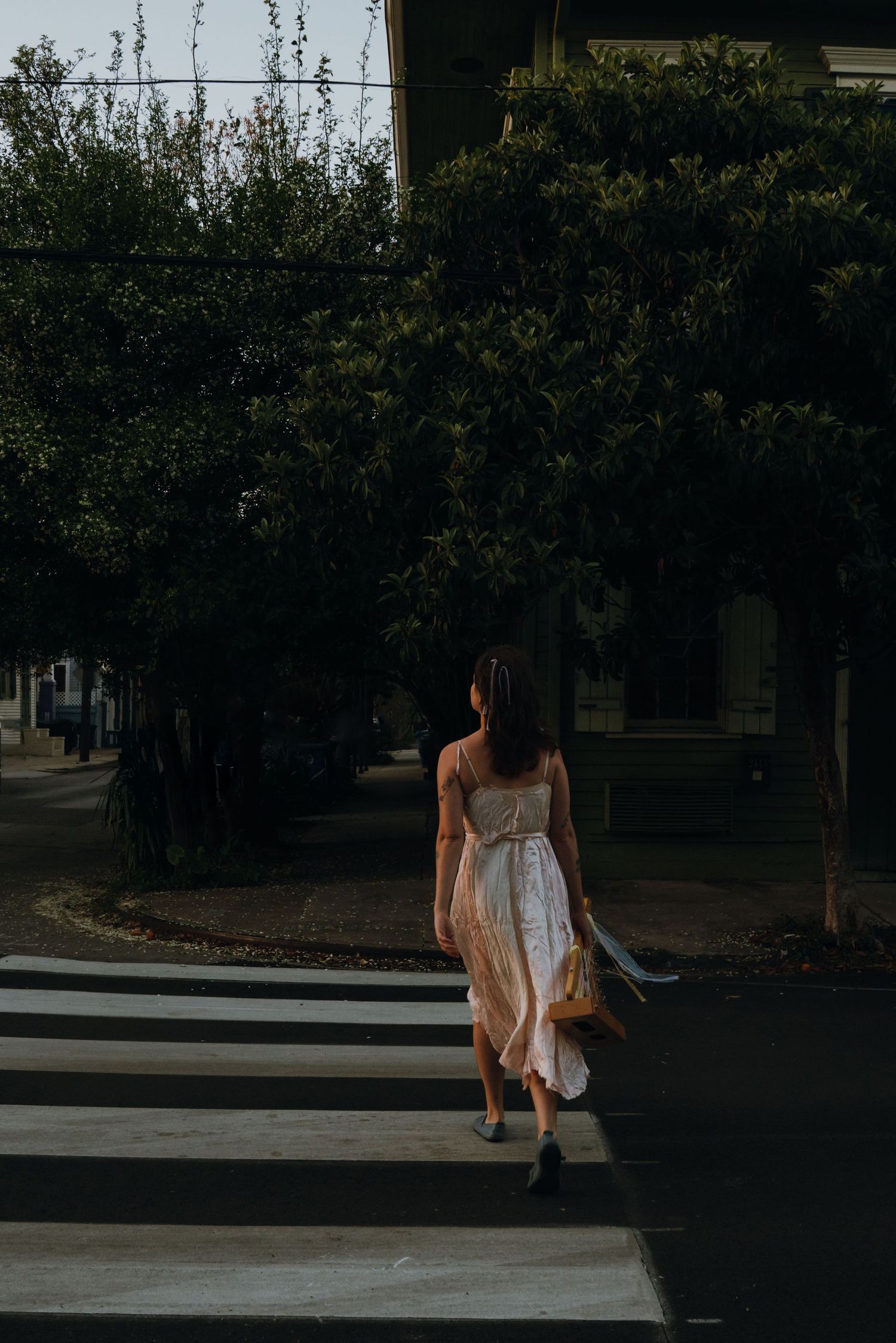 Woman in white midi dress walks across a crosswalk lined with green trees in soft evening light.