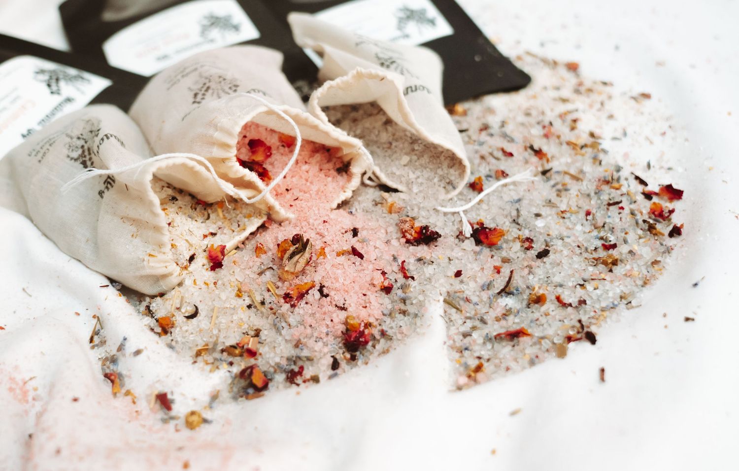 Natural bath salt sachets in muslin bags with pink and grey salts and dried botanicals scattered around.