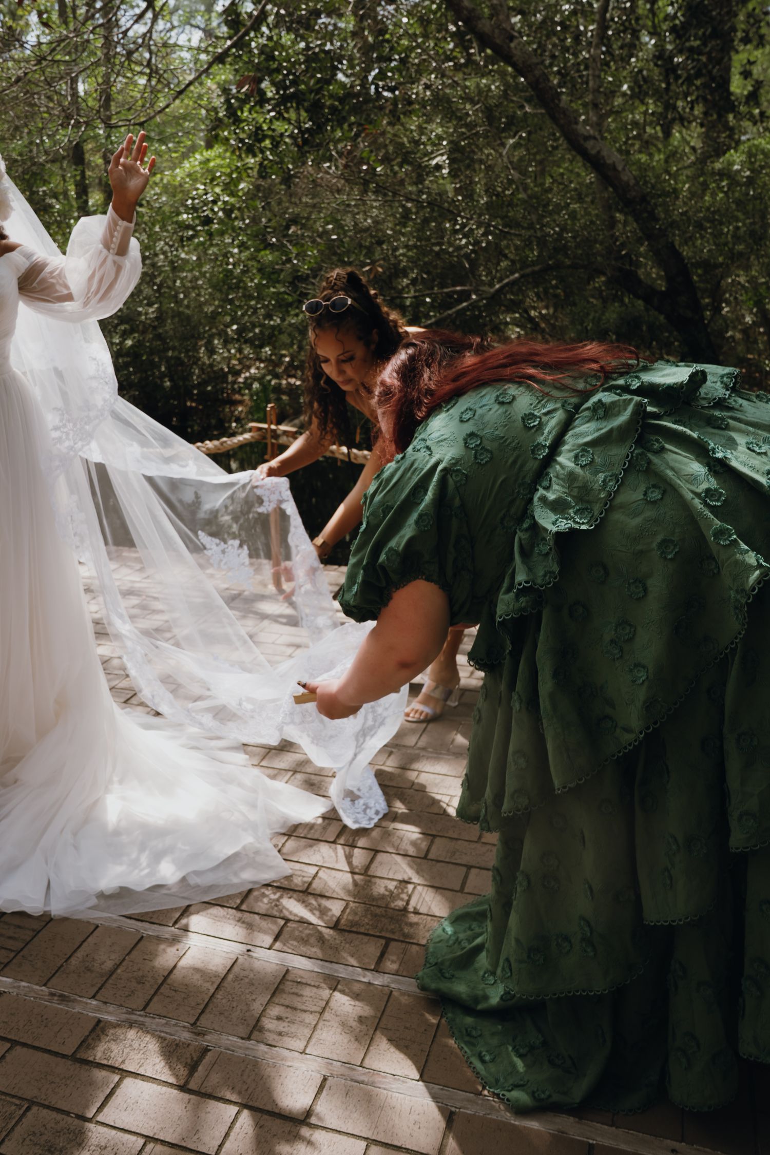 Bridesmaids in green dresses arrange bride's flowing white veil on outdoor path.