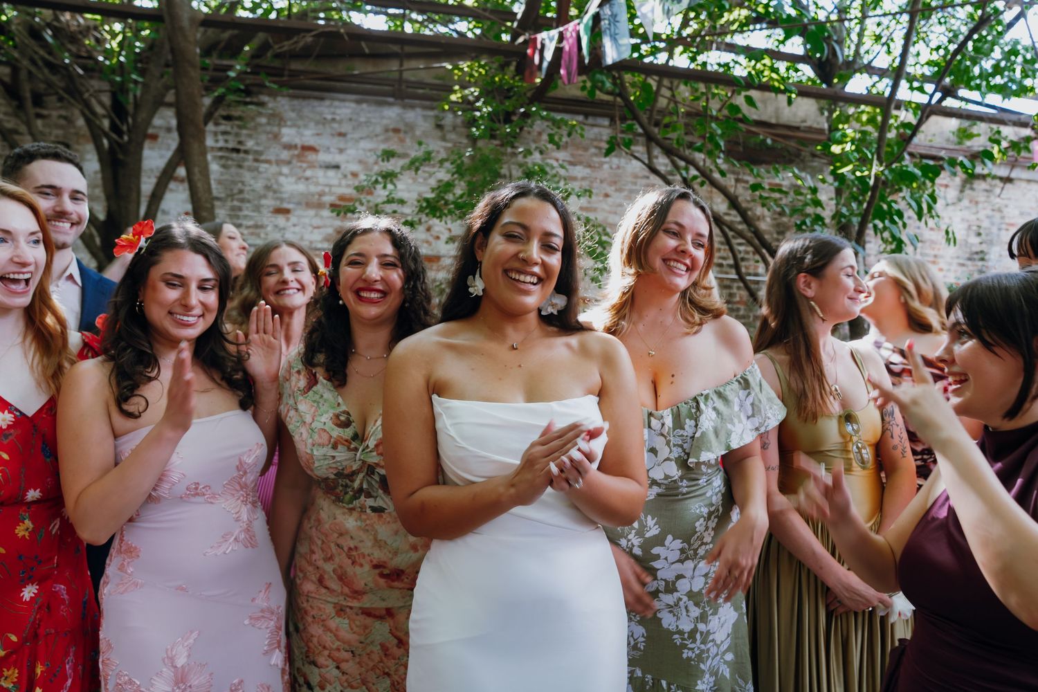 Joyful bride surrounded by smiling bridesmaids in floral dresses celebrating at outdoor wedding.
