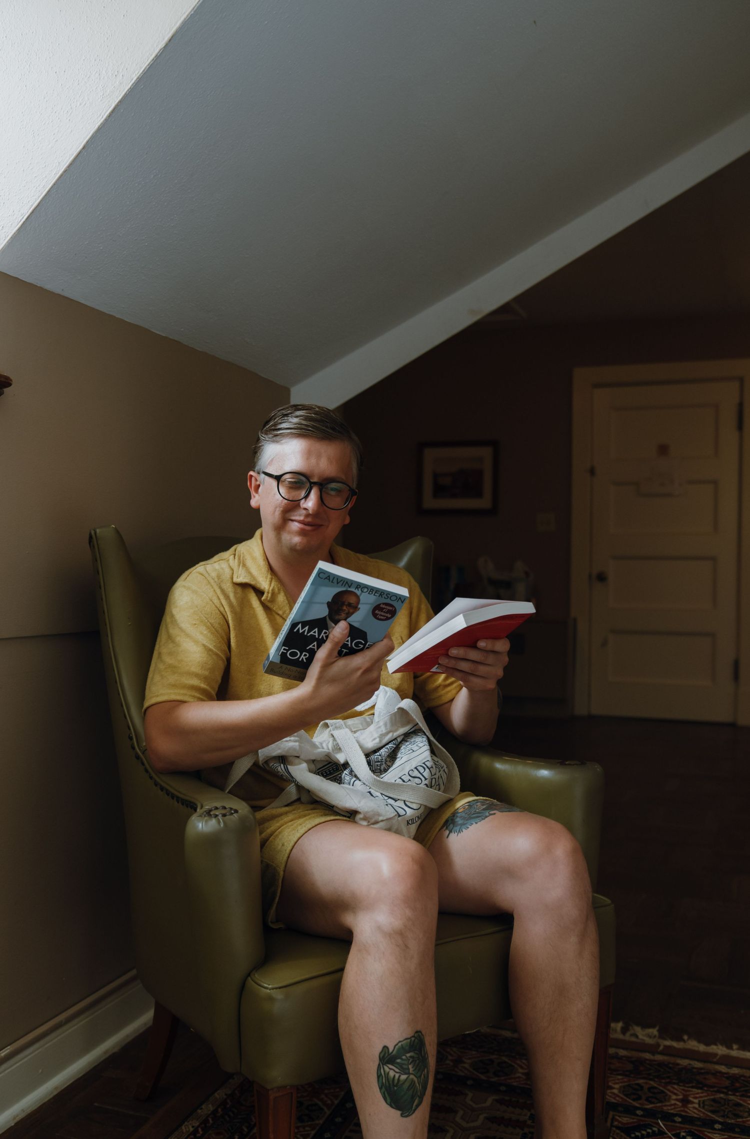 Man in yellow shirt and shorts sits reading book in green leather armchair in cozy room.
