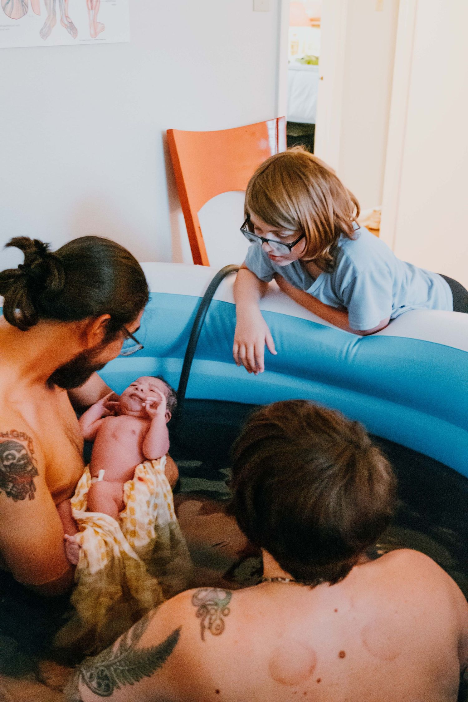 Tattooed father holds newborn baby in birthing pool as siblings look on with wonder.