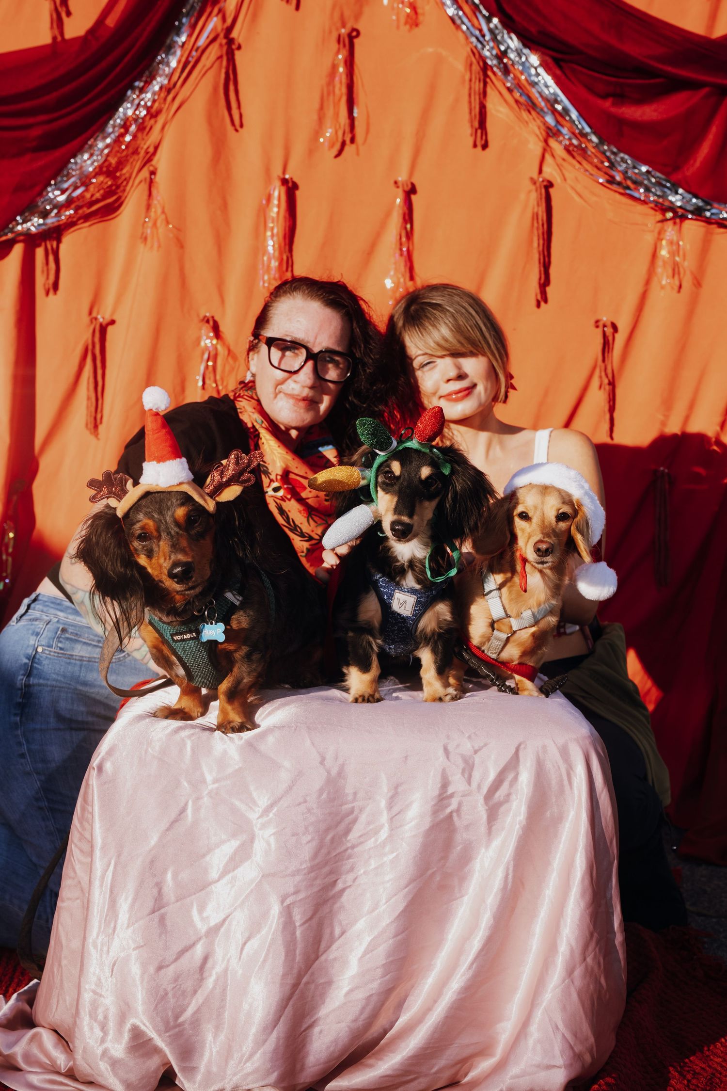 Two women pose with three dachshunds in festive Christmas outfits against a red holiday backdrop.