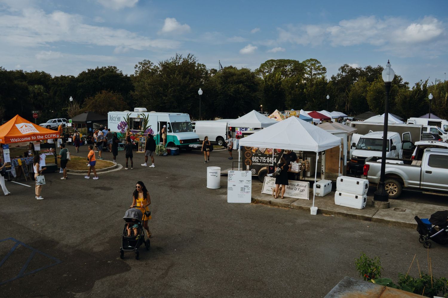 Aerial view of an outdoor market event with white tents, food trucks, vendors, and visitors in a parking lot.