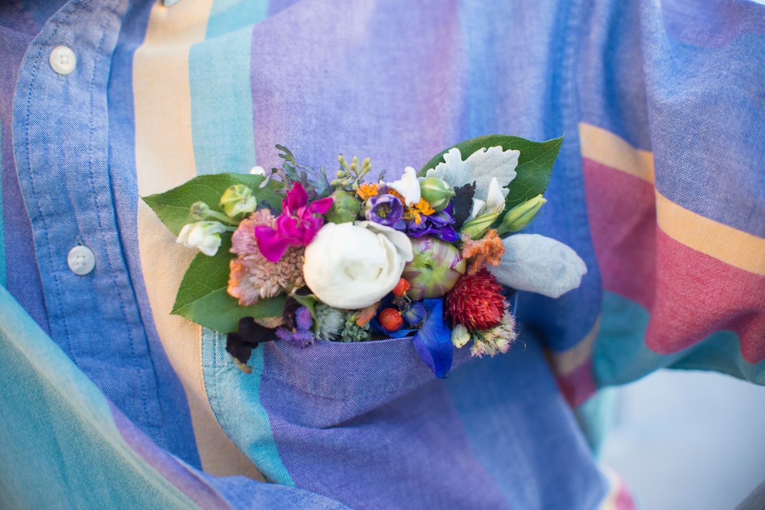 Colorful floral boutonniere with white ranunculus and mixed blooms tucked into a blue linen shirt pocket.