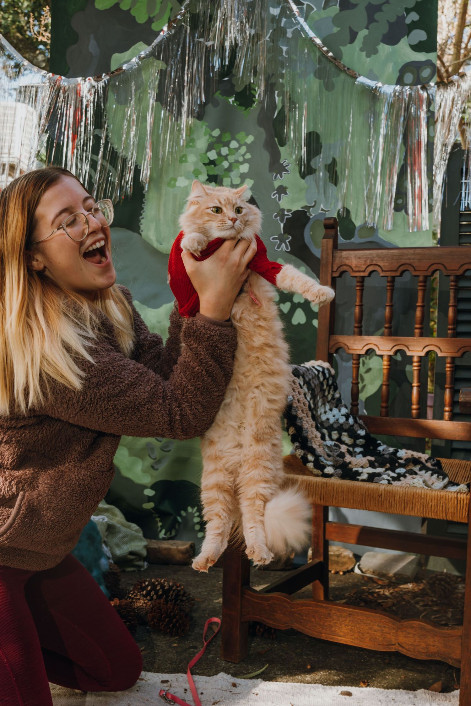 A laughing woman in glasses holds up a fluffy orange cat wearing a red outfit near a decorated holiday backdrop.