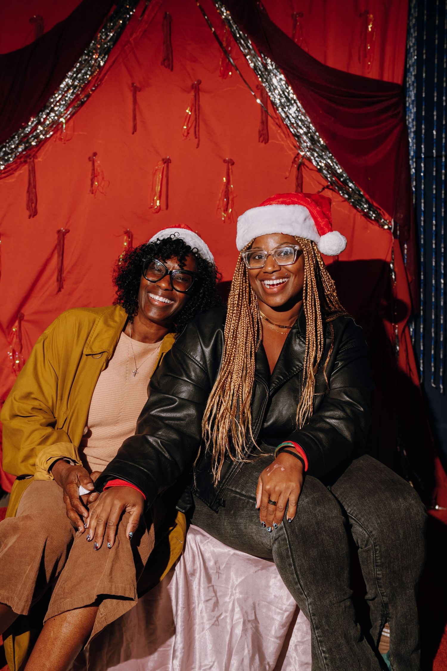 Two smiling women wearing Santa hats pose together in front of festive red curtain backdrop with silver tinsel.