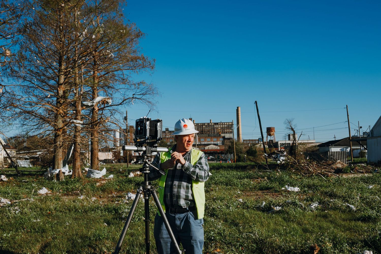 Photographer in hard hat setting up camera on tripod amid storm debris in open field.