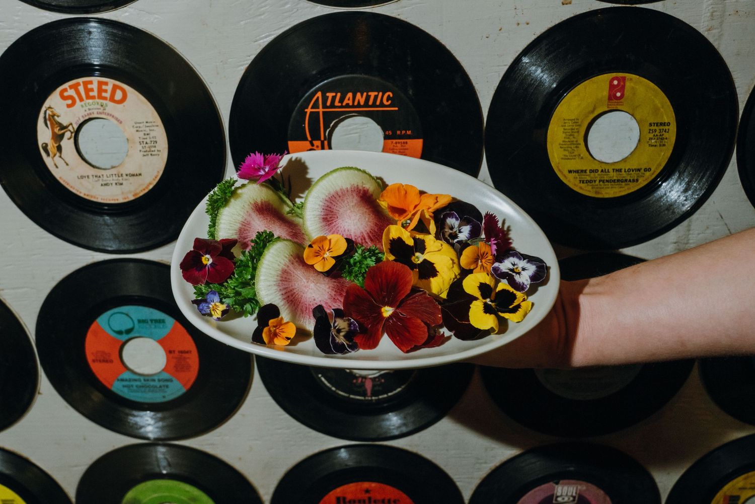 A hand holds a floral salad plate in front of a wall of vintage vinyl records including Atlantic and Steed labels.