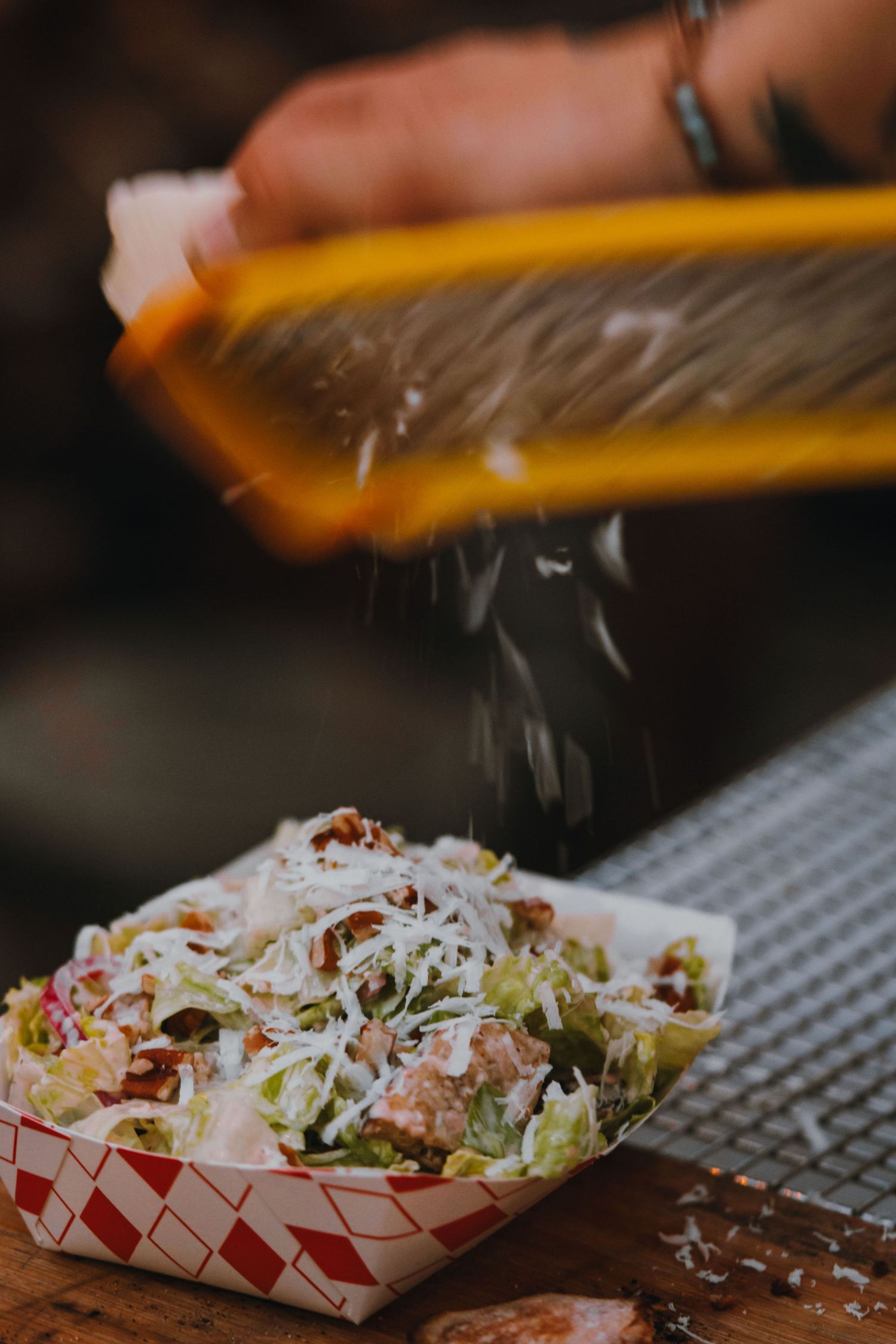Chef grating fresh parmesan cheese over a Caesar salad served in a red checkered paper tray.