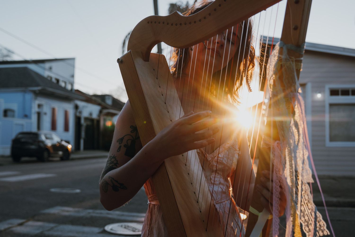 Tattooed musician playing wooden harp on street corner with sun flare at golden hour.