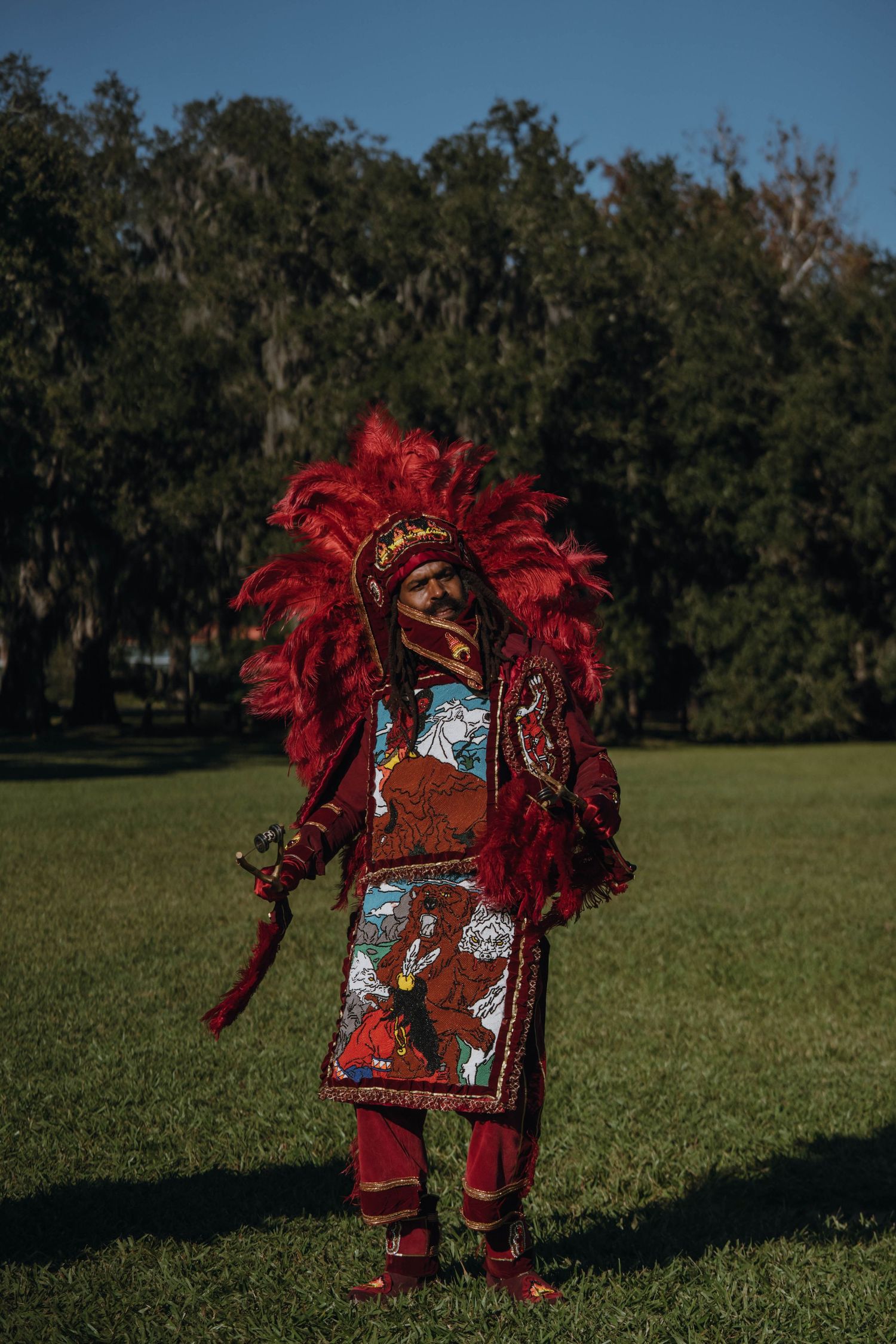 Mardi Gras Indian in full red feathered regalia with detailed beadwork panels stands in a sunny park.
