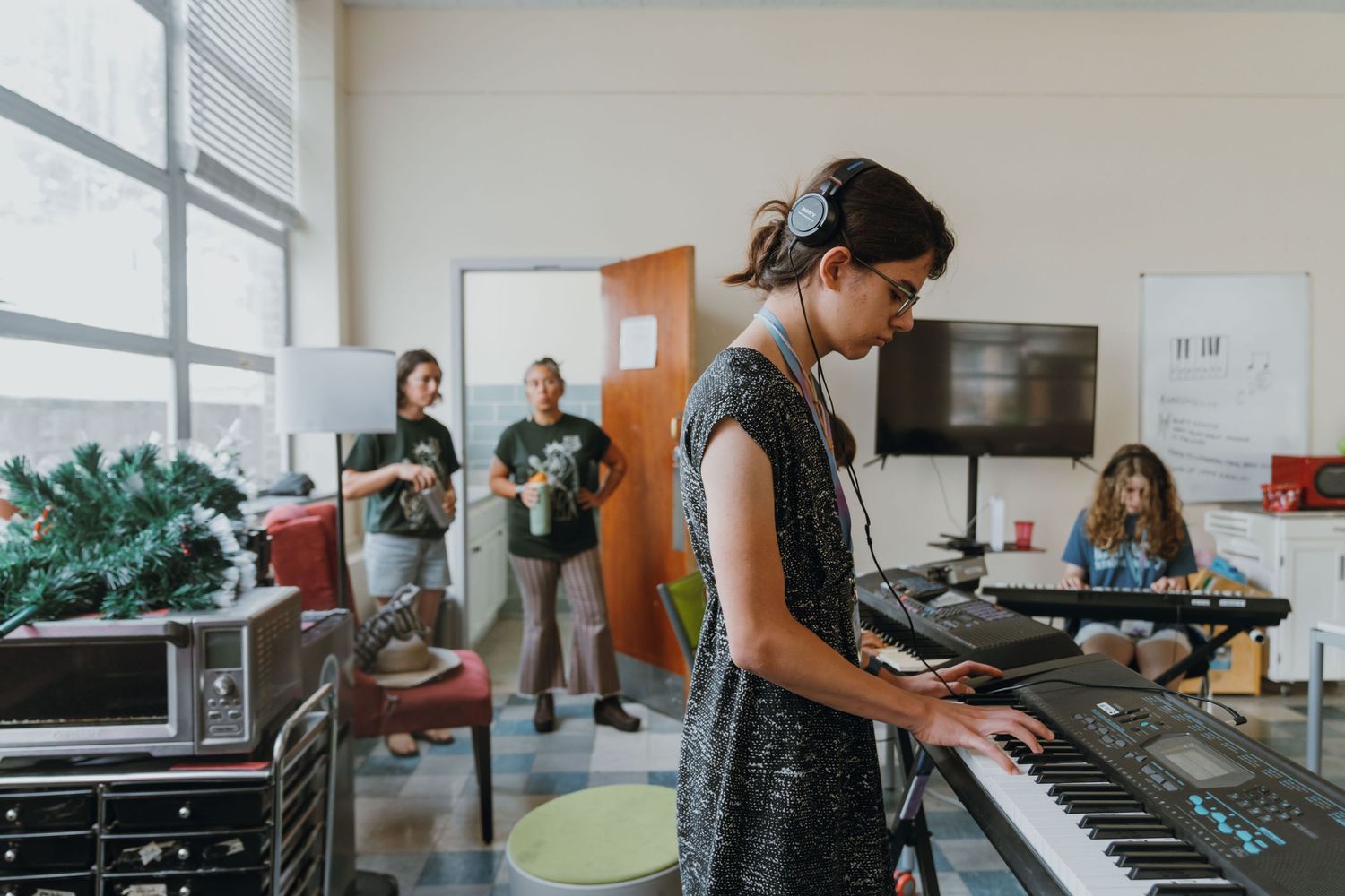 Woman with headphones plays electronic keyboard in a bright classroom while others watch in background. New Orleans, Louisiana, Mississippi