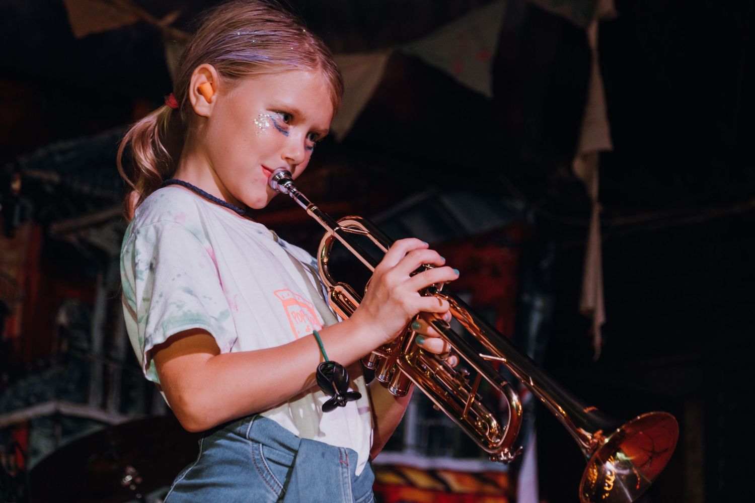 Young girl playing a brass trumpet on stage at a colorful music venue.