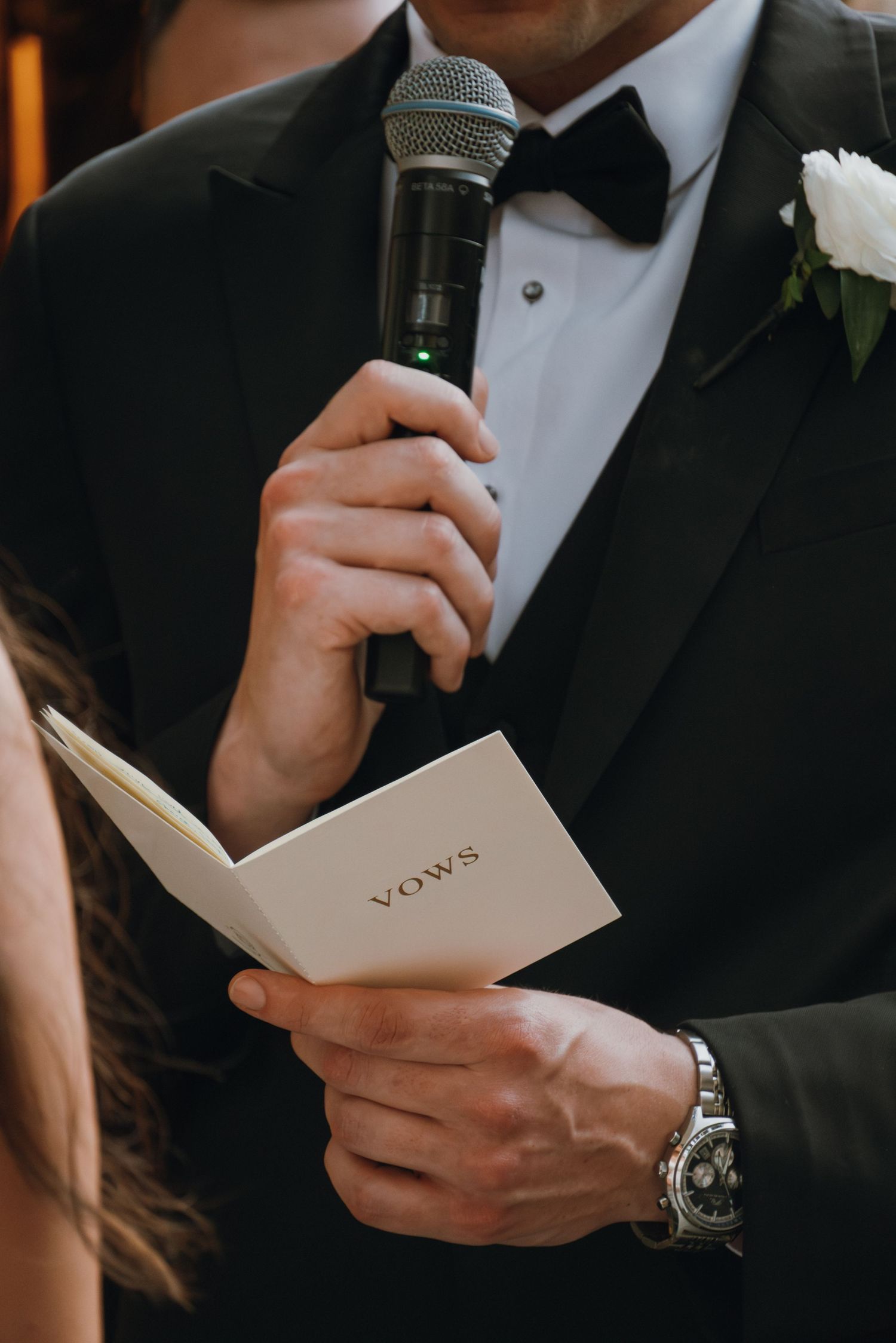 Groom in tuxedo reading wedding vows from booklet while holding microphone during ceremony.