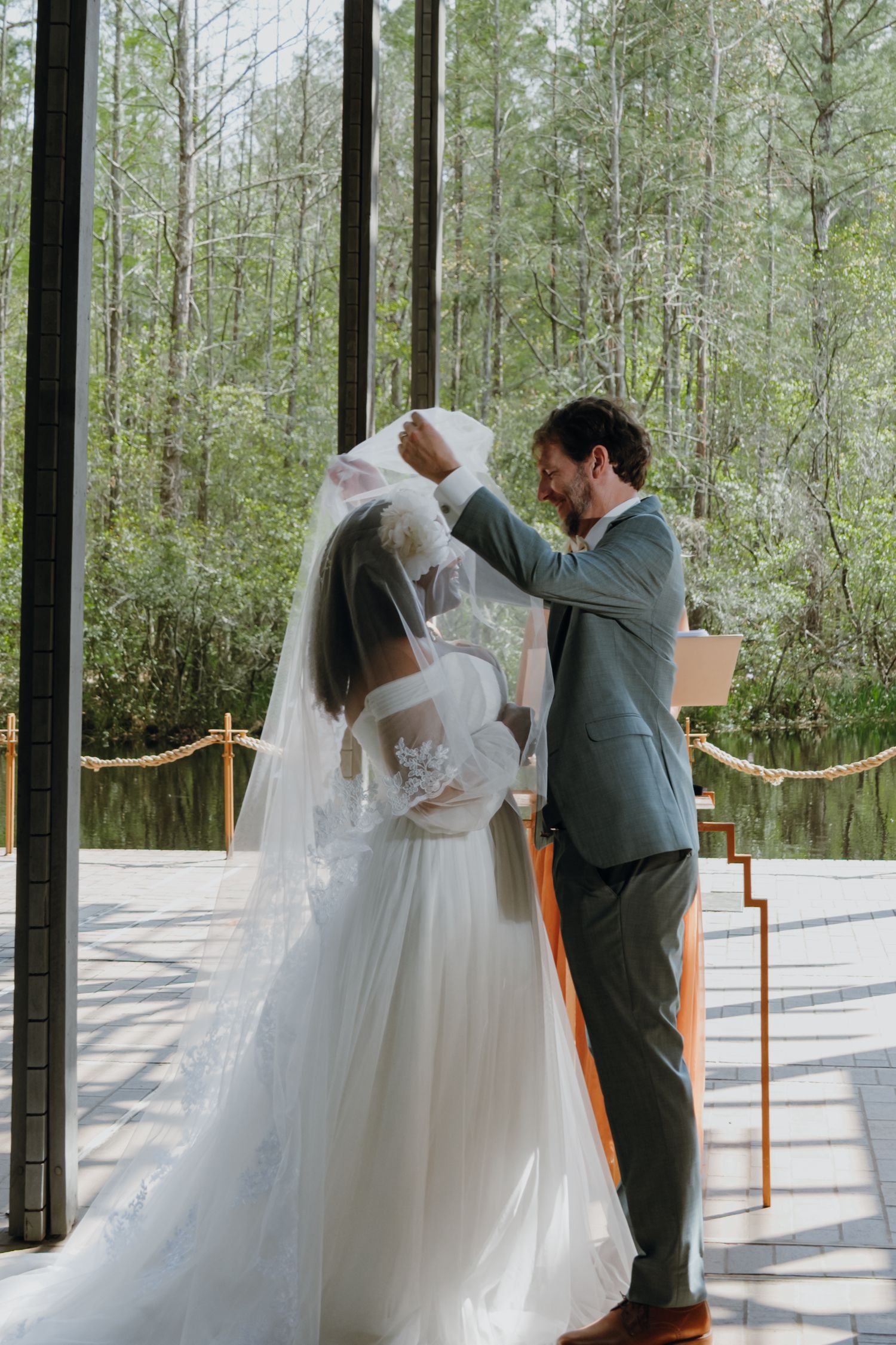 Groom tenderly lifts bride's veil during intimate wedding ceremony in glass chapel.