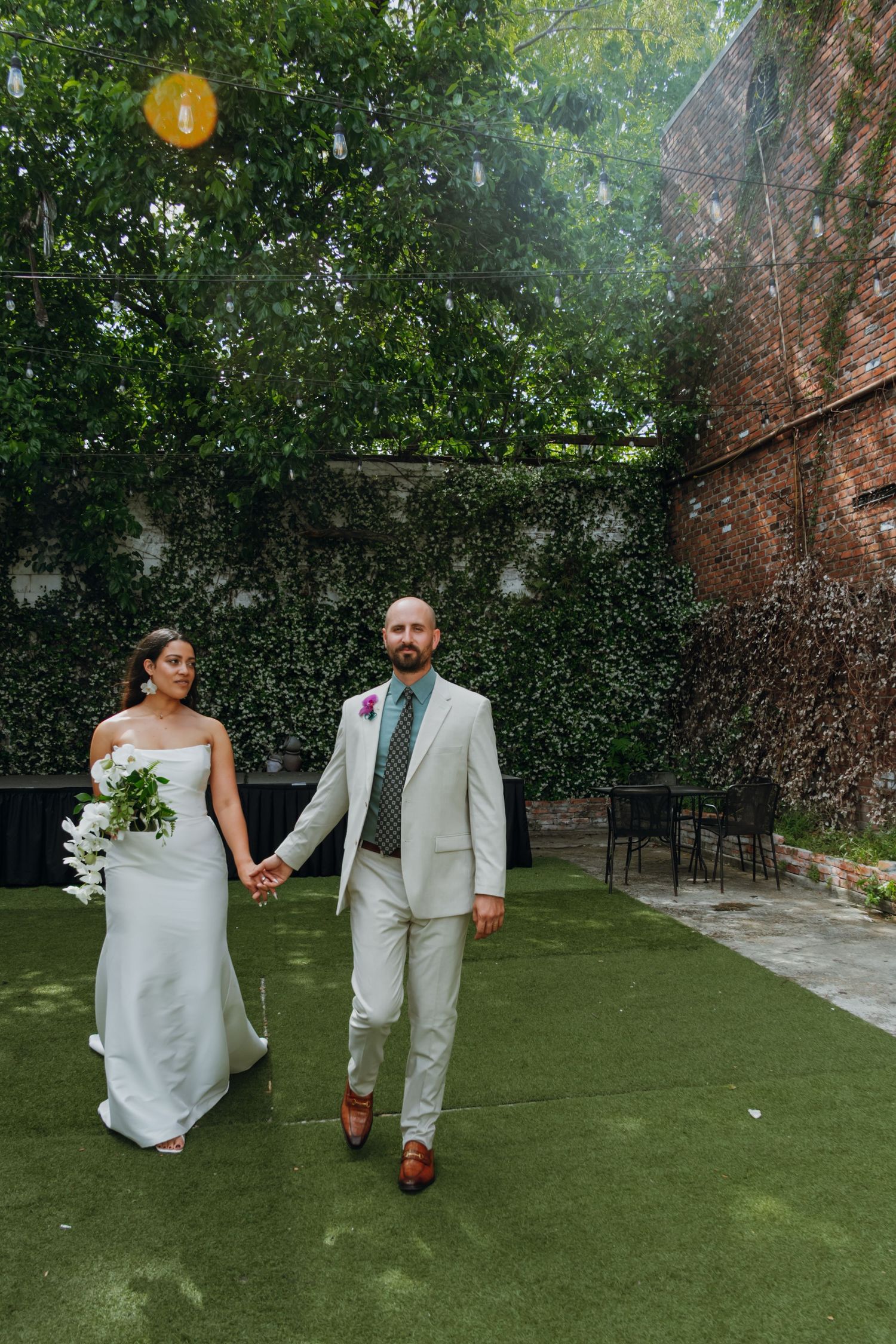 Bride and groom walk hand in hand across green lawn surrounded by lush garden walls.