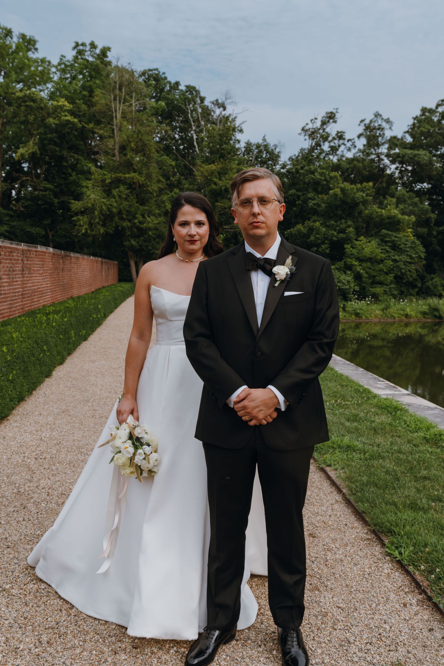Bride and groom pose on a garden pathway, bride holding white bouquet in a strapless gown beside suited groom. New Orleans, Louisiana, Missi