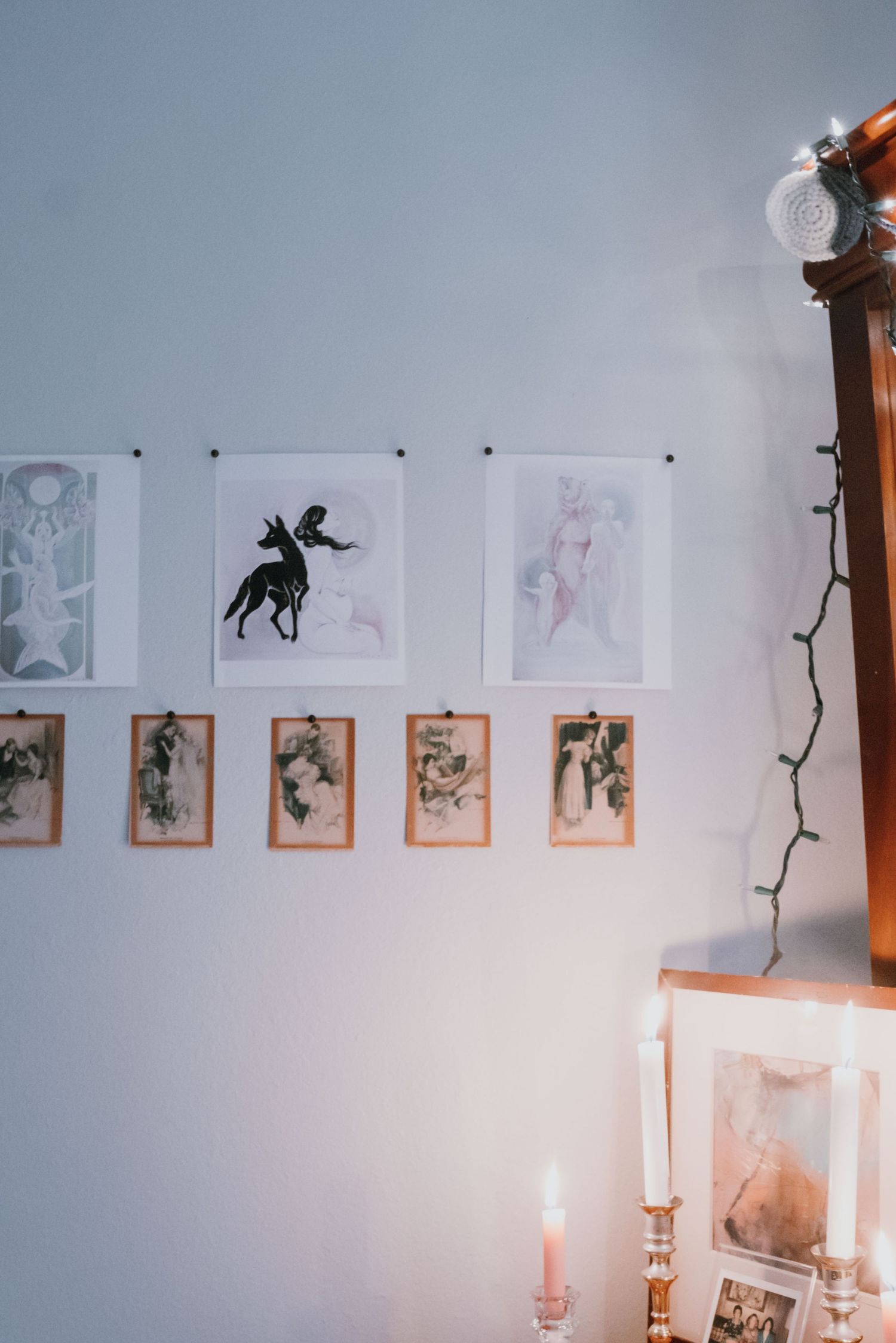 Artistic prints and illustrations displayed on a white wall above candles and fairy lights in a cozy room.