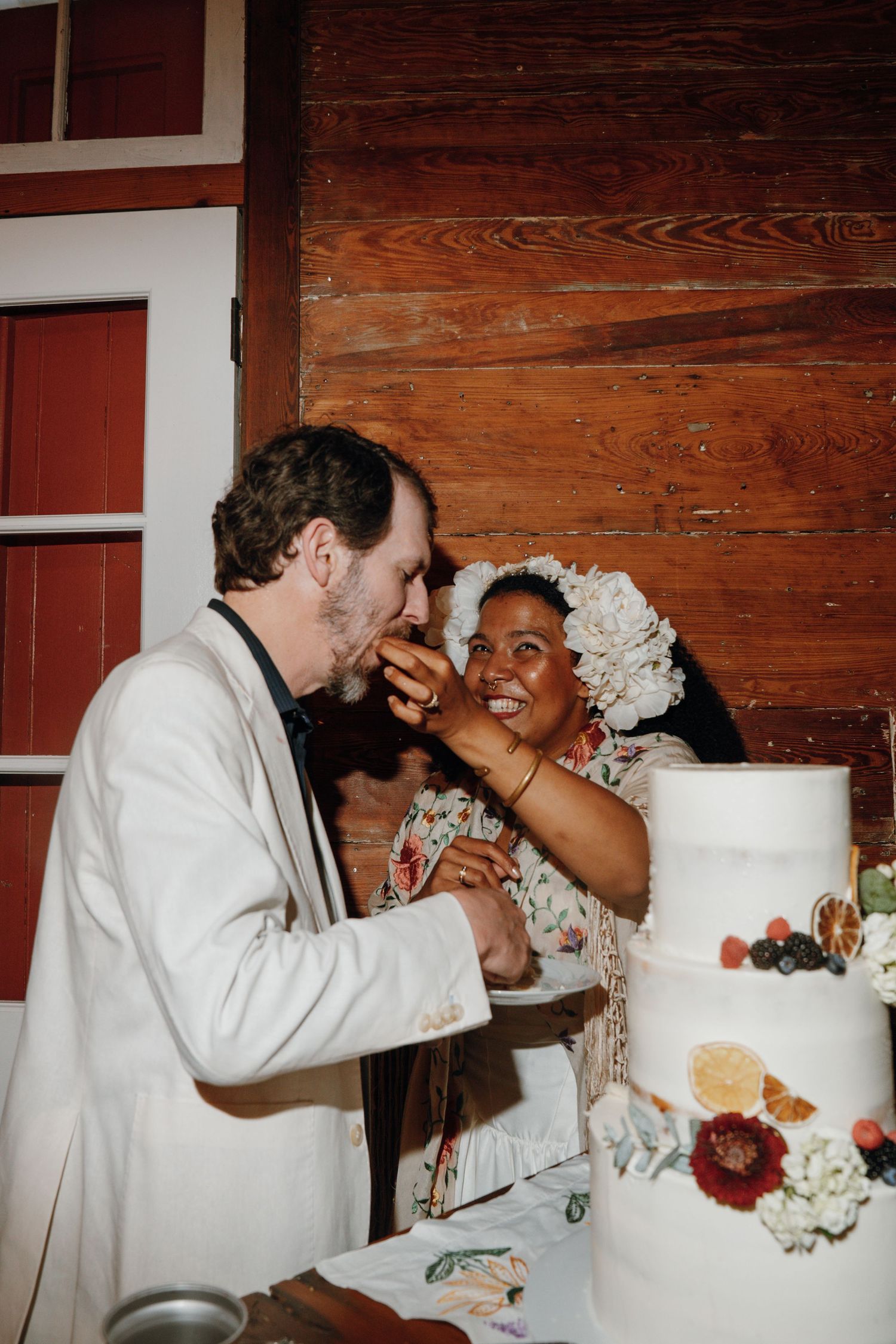 Groom feeds bride a piece of cake at their rustic wooden wedding reception venue.