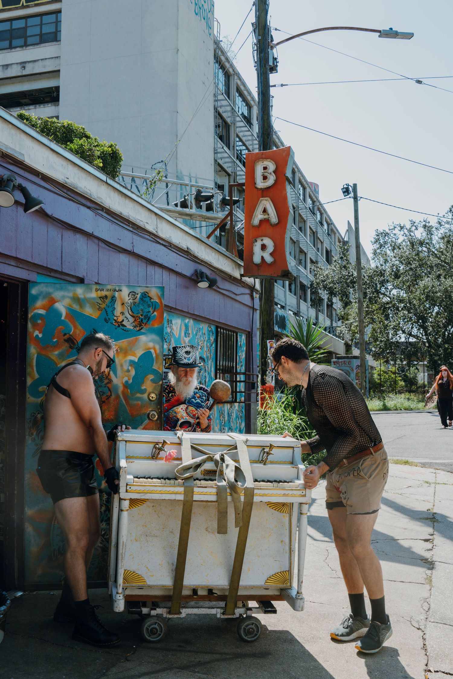 Two people unload a cart outside a bar with neon sign and colorful mural on a sunny urban street.