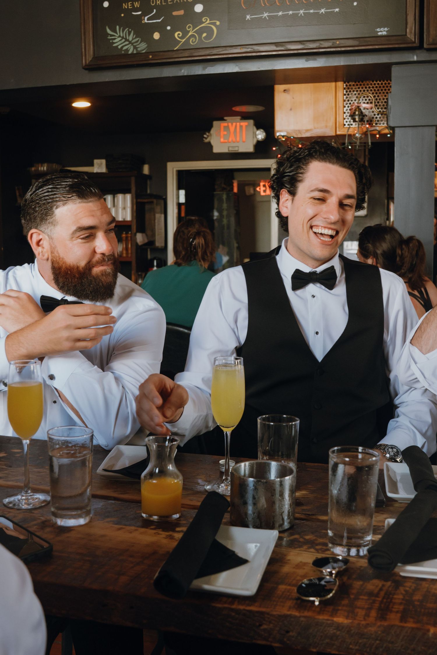 Two groomsmen laughing together at a bar table with mimosas during pre-wedding celebrations.