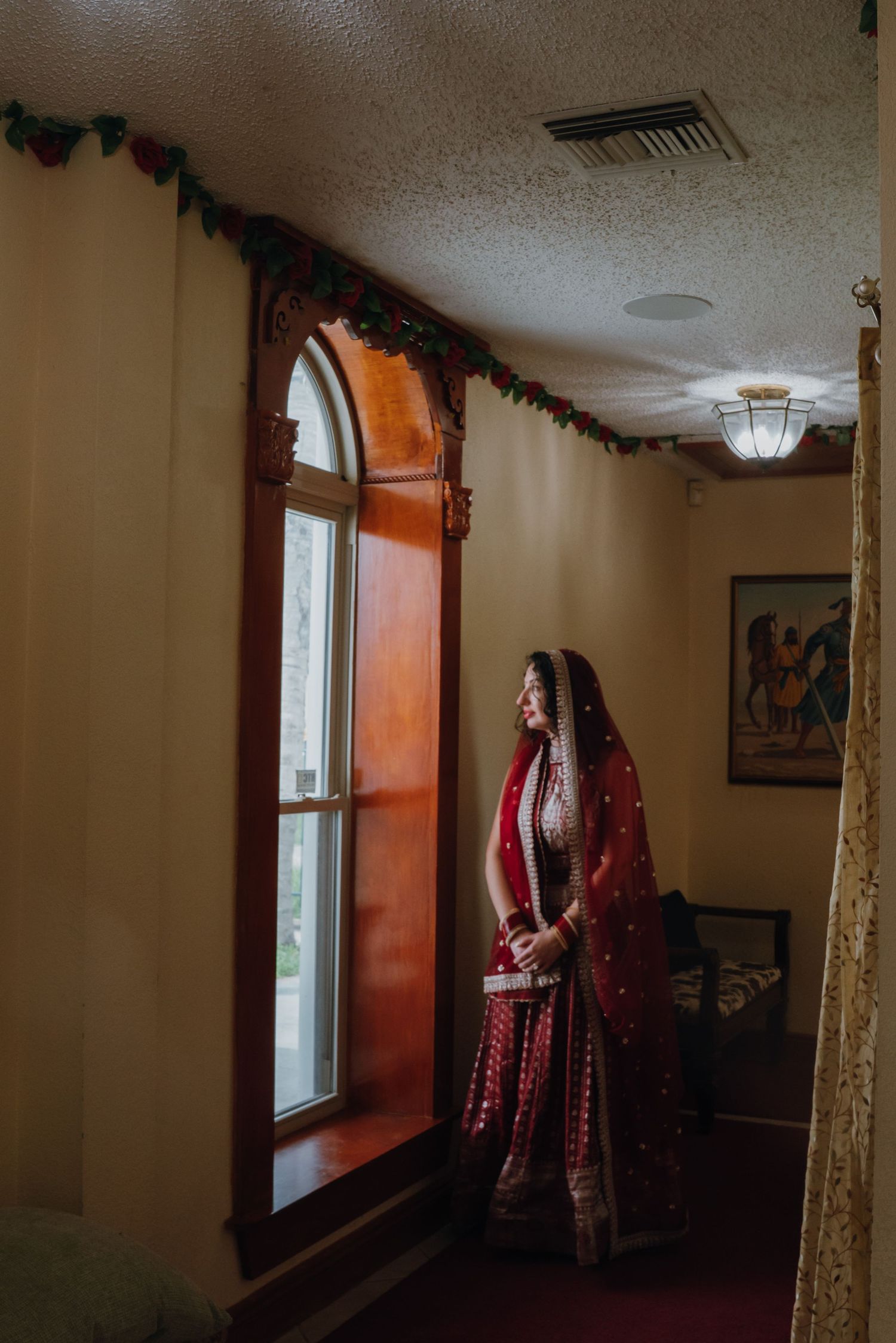 Sikh bride in stunning red lehenga with dupatta stands by an arched window in soft natural light.
