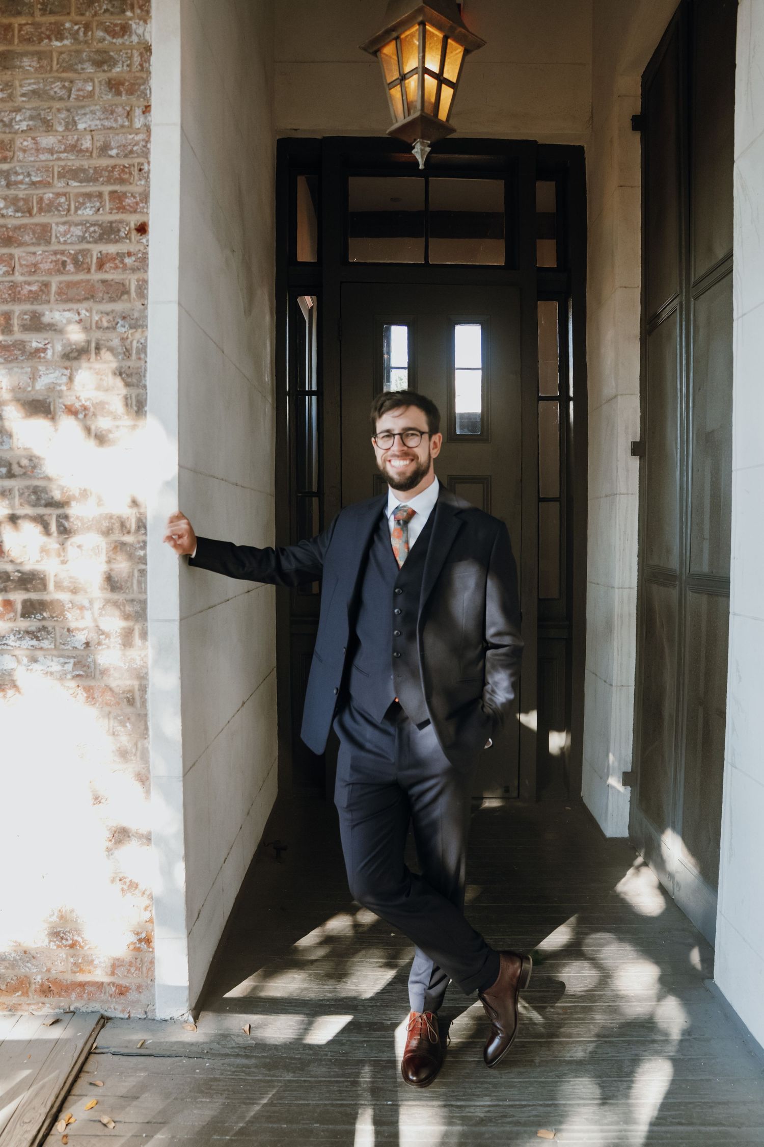 Smiling groom in navy suit leaning against wall by elegant dark door with lantern.