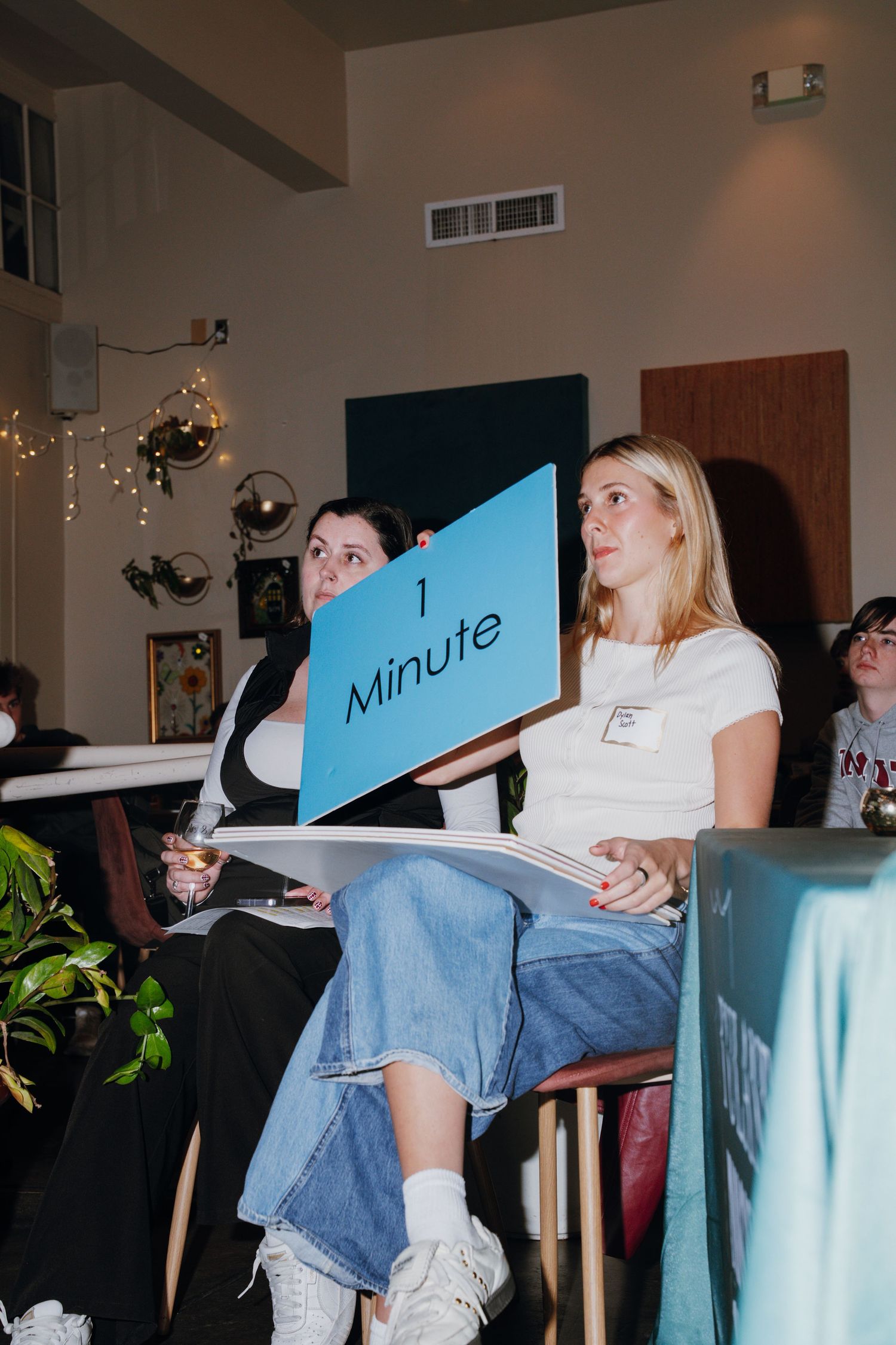 Woman holding blue '1 Minute' sign at indoor event, blonde woman seated beside her.