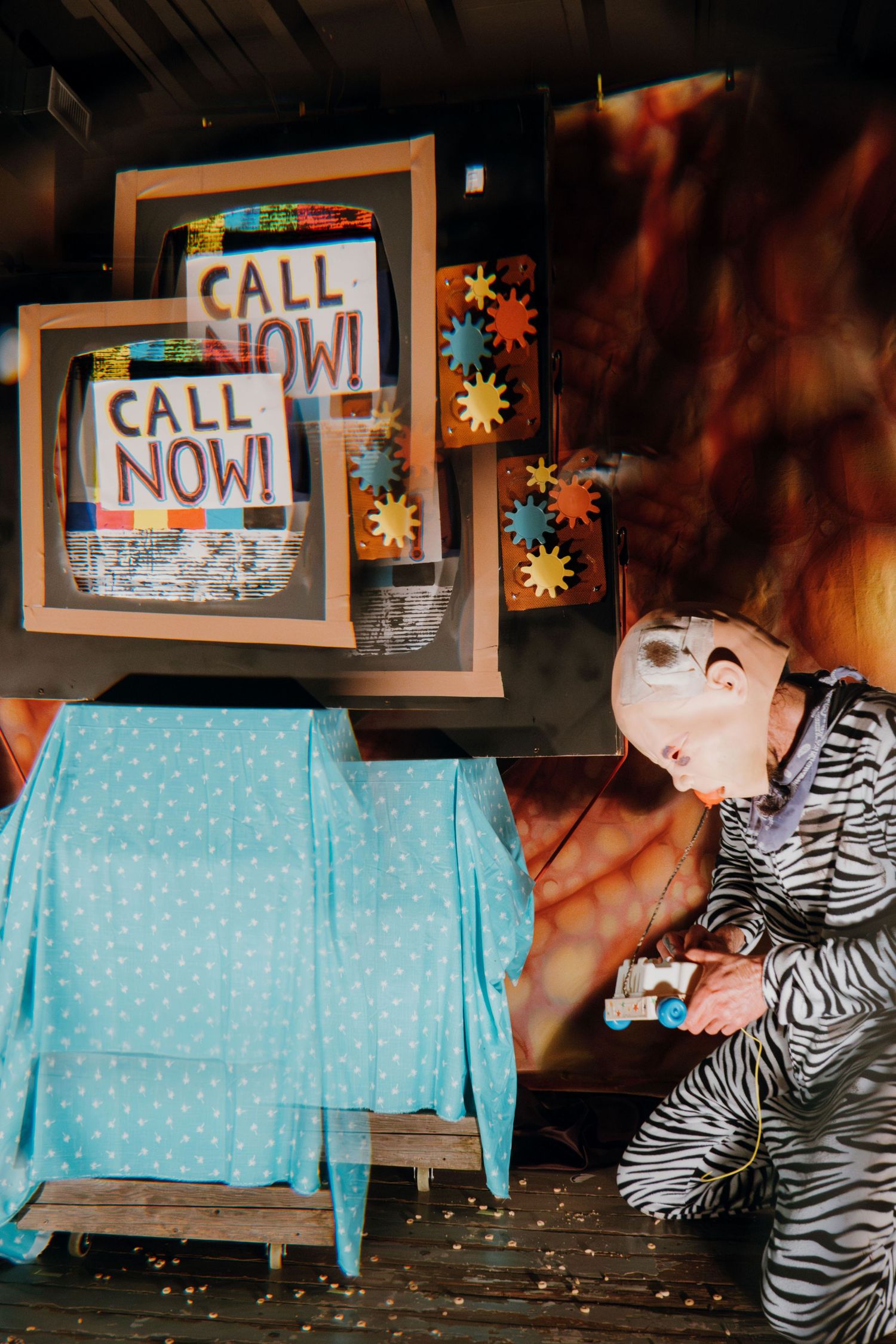 Costumed performer crouches near Call Now signs in quirky theatrical stage set.