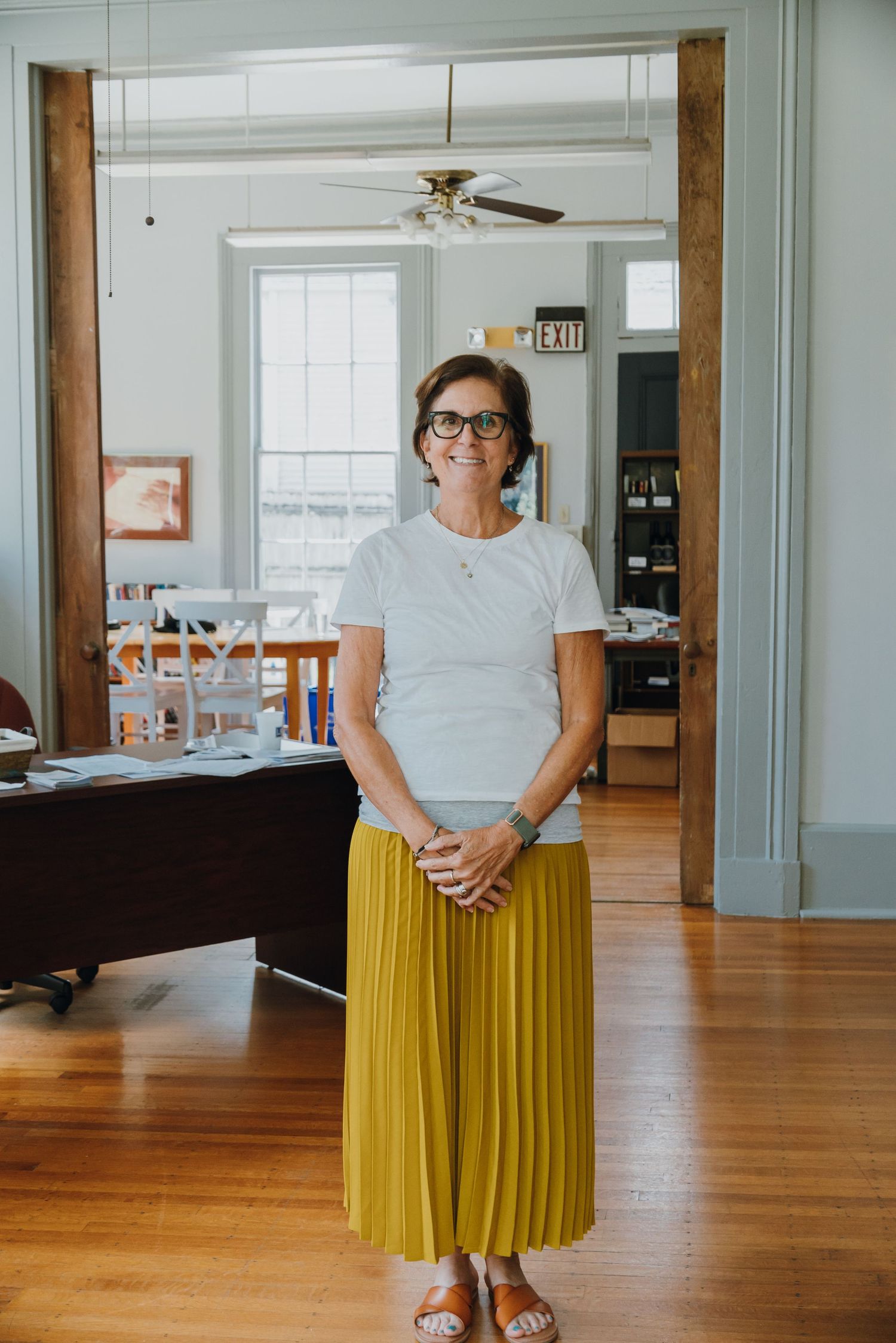 Smiling woman in white top and yellow pleated skirt standing in a bright wooden-floored office space.