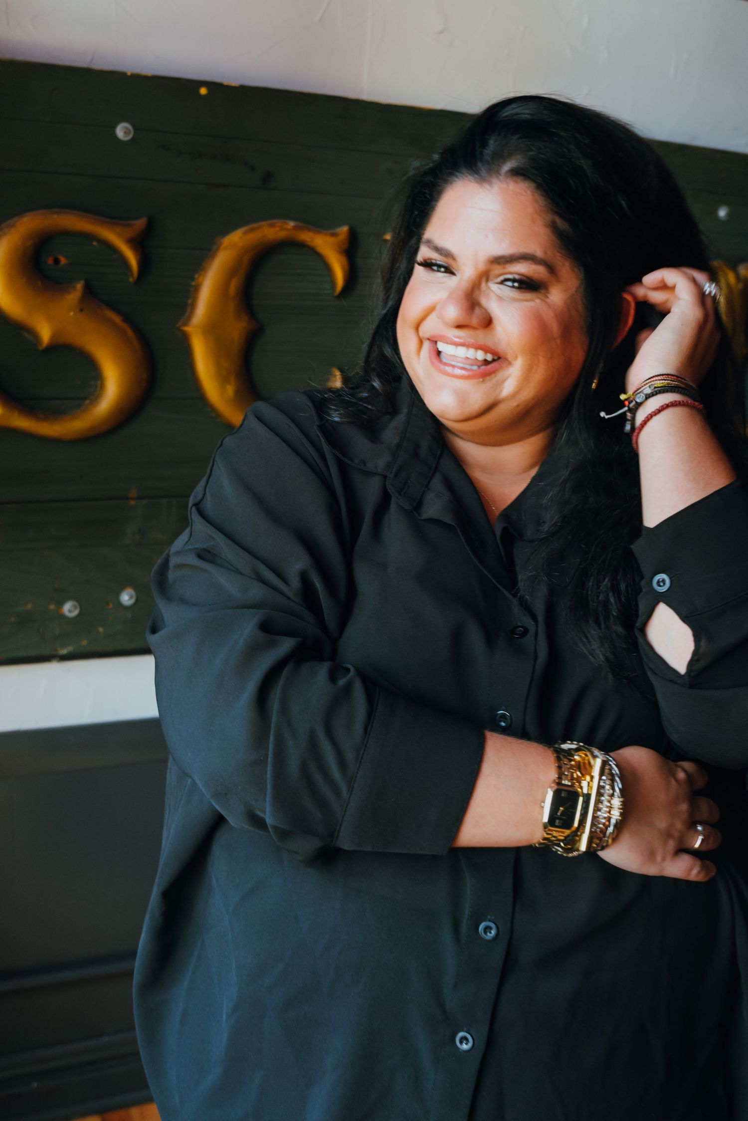 Cheerful woman in black shirt laughing while touching her hair near a dark green vintage sign.