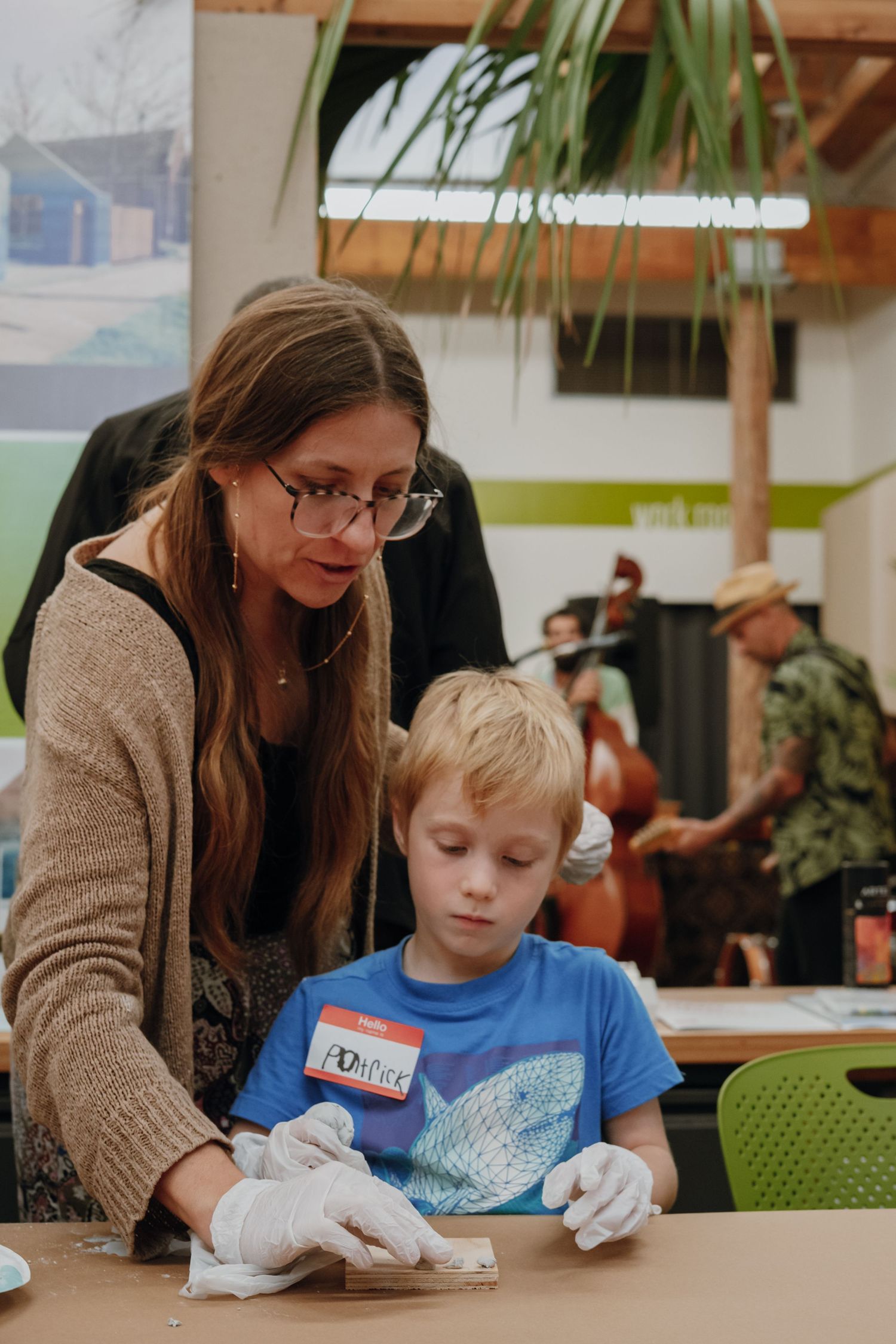 Woman helps young boy with craft activity at community event while live music plays in background.