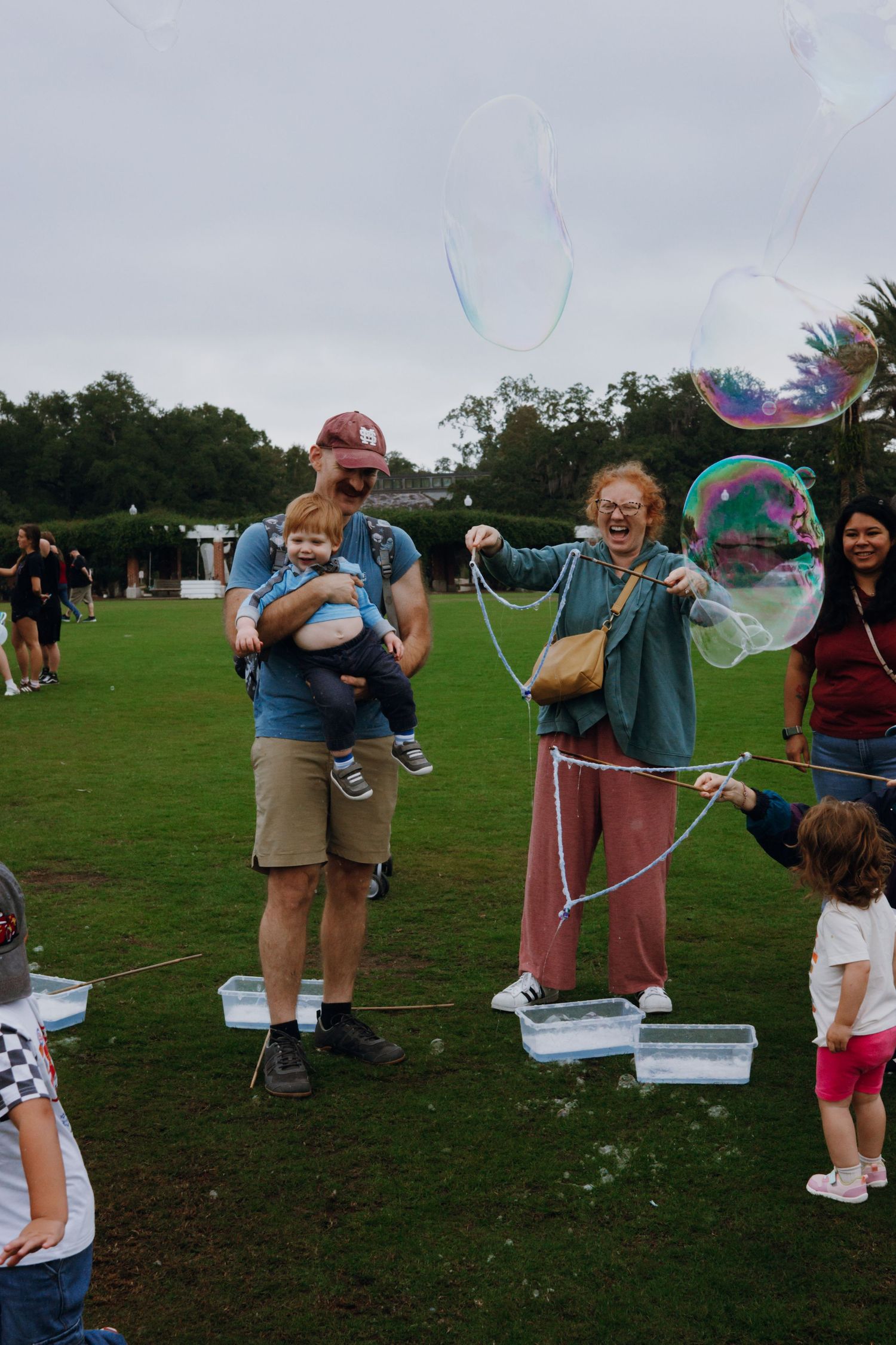 Family and children playing with giant soap bubbles outdoors at a park event on a cloudy day.