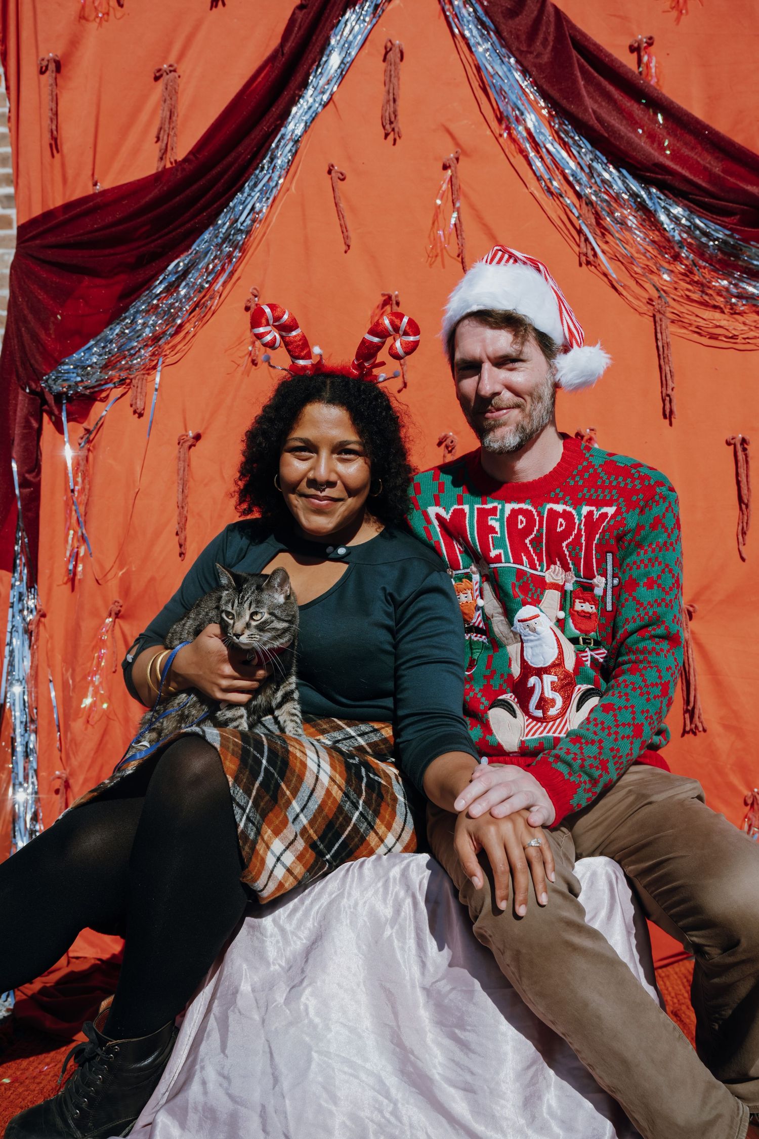 Couple in Christmas sweater and reindeer headband pose with tabby cat before festive red backdrop.