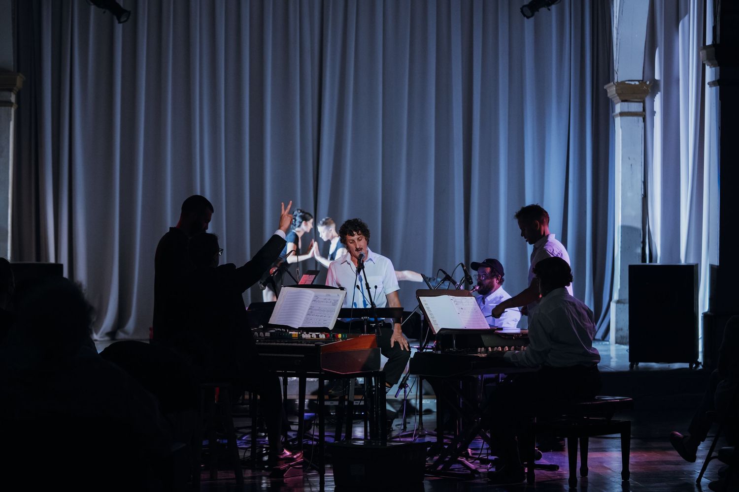 Musicians perform on stage with music stands under dramatic lighting against a white curtain backdrop.