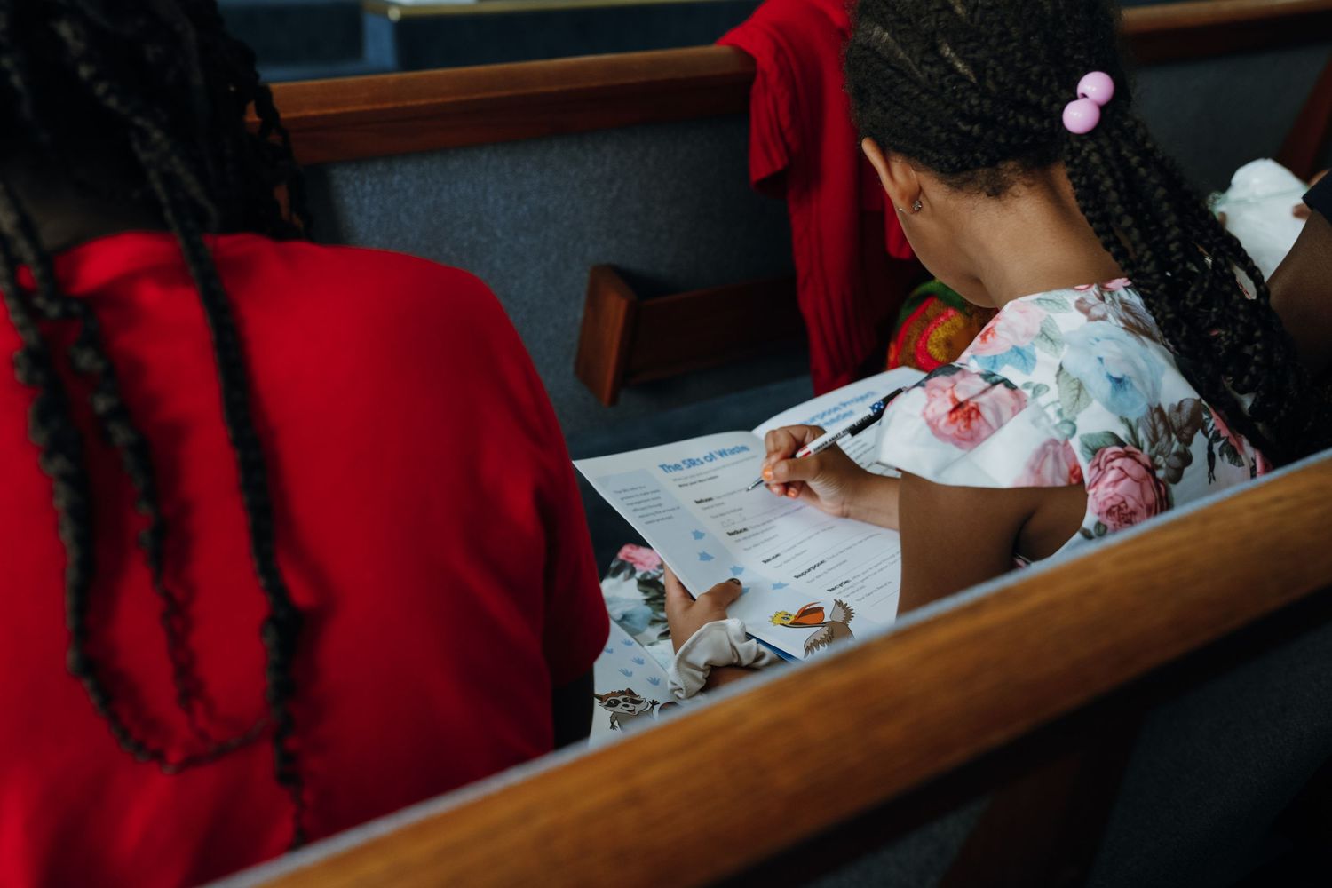 Young girl with braids writing on paper in church pew beside woman in red dress.